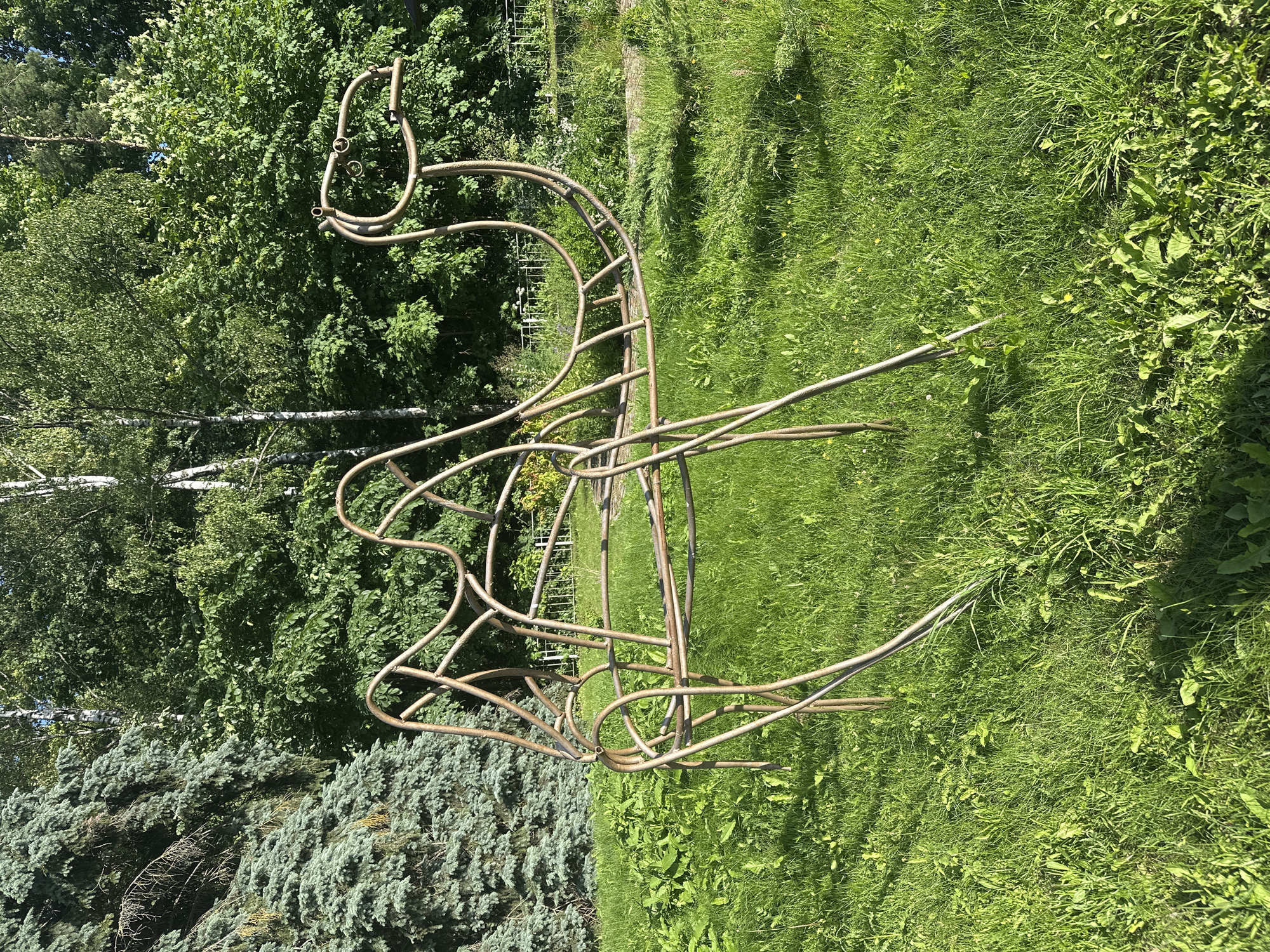 A metallic garden sculpture resembling a flamingo stands in a grassy yard. The sculpture is made of thin, curved metal tubes and has an elongated neck, a curved body, and two legs extending downwards. The flamingo's head is formed by a loop with two smaller loops attached, suggesting eyes. The sculpture is a muted metallic color, possibly brass or weathered steel.
The scene is set in a bright, outdoor environment with lush green grass and trees in the background. To the left of the sculpture, a dense cluster of evergreen trees with blue-green foliage is visible. Behind that, a mix of deciduous trees with vibrant green leaves and slender white tree trunks suggests a mixed woodland. The grass appears to be a natural lawn, with some taller weeds and wildflowers interspersed.
The lighting indicates it is daytime, likely sunny, as evidenced by the clear shadows and bright illumination of the scene. The overall mood is peaceful and tranquil, typical of a garden setting on a pleasant day. There is no visible text in the image. The image appears to be taken from a slightly elevated angle, looking down at the sculpture and its surroundings.