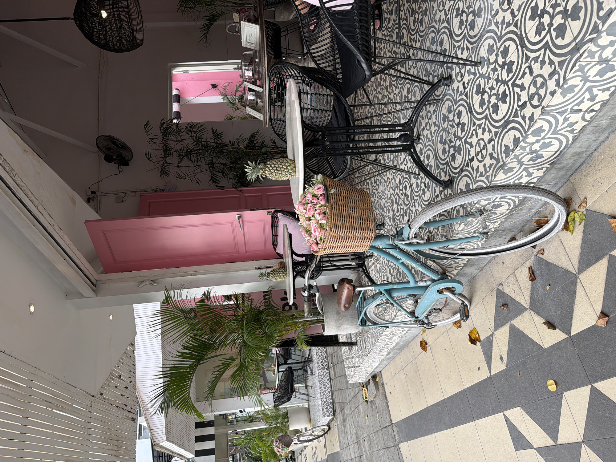 The scene depicts the exterior of a café in Kuta, Indonesia, marked by a visually distinctive aesthetic. A light-blue bicycle, adorned with a flower-filled basket, stands prominently near the café's seating area. Tables and black woven chairs are positioned on a patterned tile floor that transitions to a gray and white triangular tiled sidewalk. A pink door frame and pink wall section are noticeable architectural features. Tropical plants, including palm trees and other greenery, are strategically placed, contributing to the ambiance. The photograph, taken outdoors, features a lamp, and provides a partial view of the café's interior through a window, along with a portion of the exterior facade.