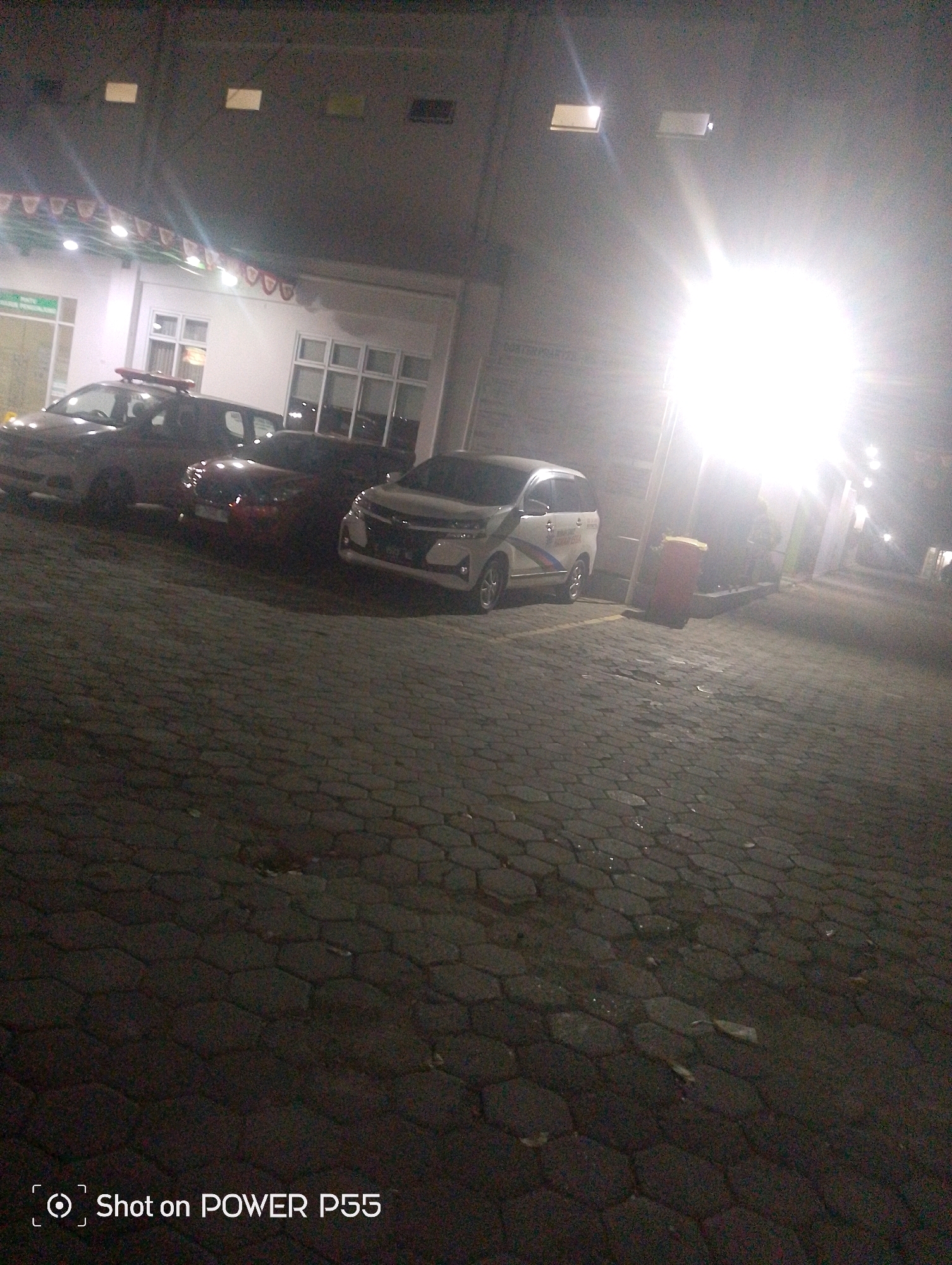 At night, a parking lot paved with hexagonal interlocking concrete blocks is illuminated by bright overhead lights. Several vehicles are parked in the lot, including a white minivan with red, blue, and yellow stripes on its side, a dark-colored SUV with a red light bar on its roof, a red car, and another white vehicle. A red trash bin is situated near the white minivan. In the background, a building with large windows and awnings is visible. The building appears to be illuminated from within, and decorative banners hang above the entrance. The scene suggests a public or commercial space, possibly a hospital or a government building, in Lebaksiu, Indonesia. The "Shot on POWER P55" watermark indicates the image was taken with a specific mobile device.