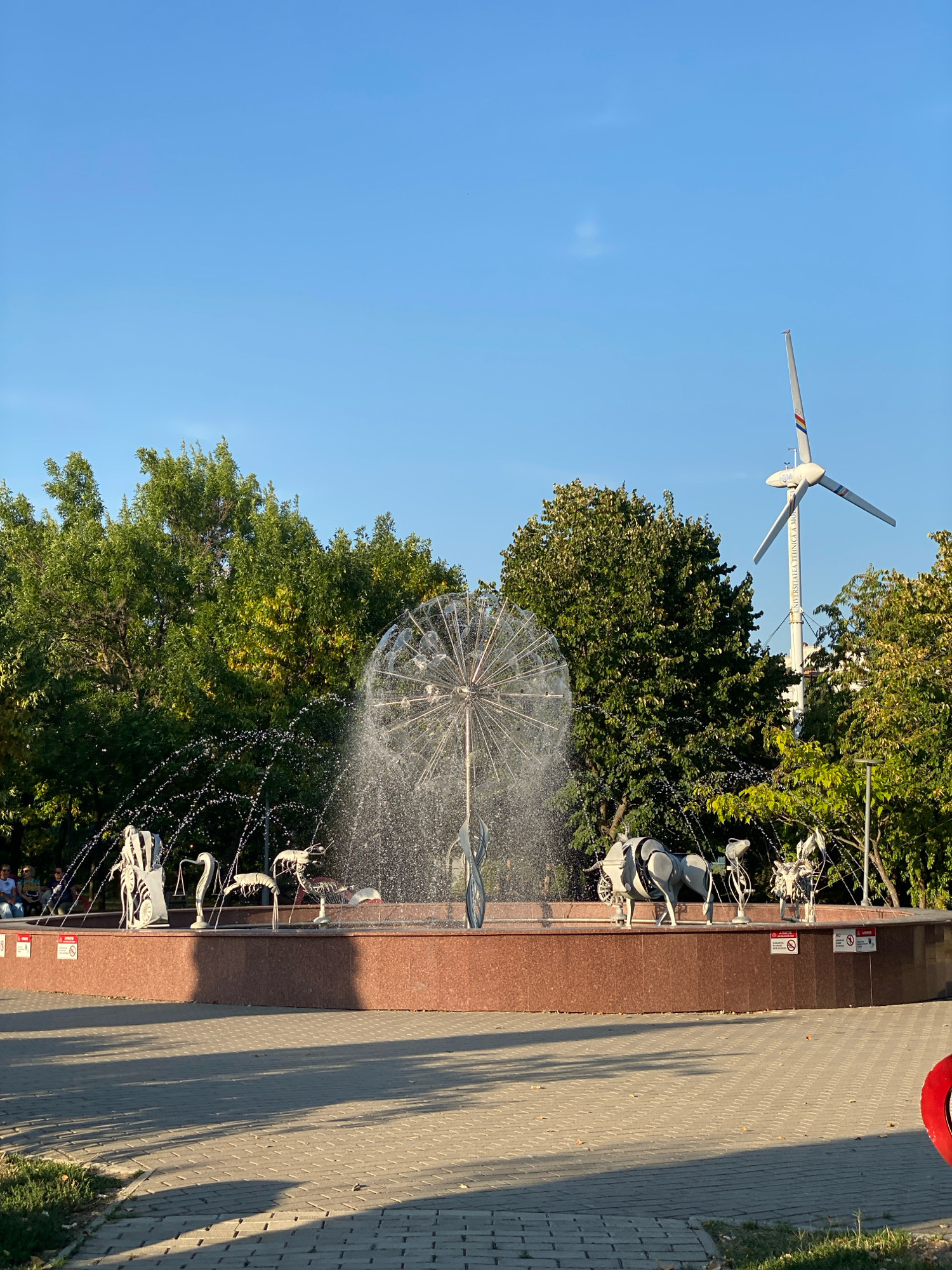 The image captures a public fountain with various animal sculptures and a wind turbine in the background. The fountain, a prominent feature, has a circular basin with a red granite exterior. Inside the basin, water sprays from a central, spherical structure, creating a misty effect. Several metal sculptures of animals, including what appears to be a seahorse, a shrimp, a bull, and possibly an elephant and a horse, are arranged around the fountain's edge. In the background, lush green trees form a dense canopy, and a tall white wind turbine with three blades stands against a clear blue sky. The ground in front of the fountain is paved with light-colored interlocking bricks, and shadows suggest it's a sunny day. A few people are seated on a bench in the distance, to the left of the fountain. The location is the University of Agricultural Sciences and Veterinary Medicine of Moldova in Chișinău, Moldova.