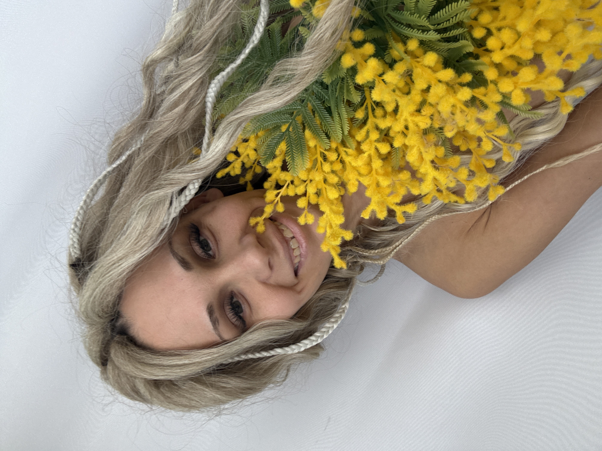 A woman with long blonde hair and a bouquet of yellow flowers is smiling at the camera. She appears to be lying on her side, with a white background. The details about location are not provided in the image.