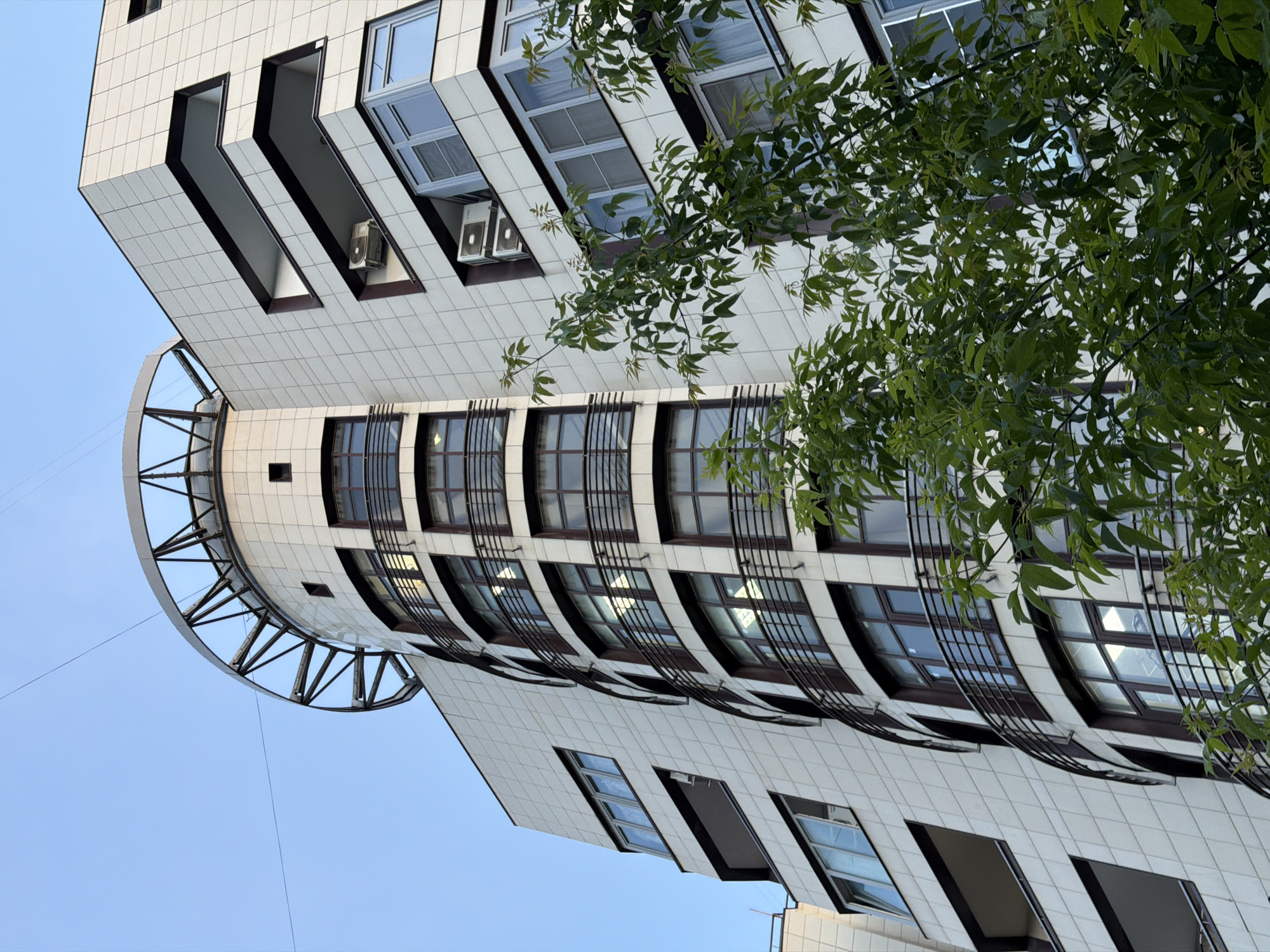 The image presents an upward, angled view of a striking modern building, partially framed by the lush green foliage of a tree on the right. The building's facade is clad in light-colored, possibly cream or off-white, square tiles with dark grid lines, creating a distinct pattern.
Architecturally, the building exhibits a dynamic and unconventional design. The upper section features several deeply recessed rectangular openings, which appear to be either balconies or very deep window frames, some equipped with external air conditioning units. Below these, the building transitions into a prominent curved section with a series of multi-paned windows set in dark frames, many reflecting the blue sky. Below this curved section, there are more levels of tiled facade with standard windows and additional recessed openings. A unique structural element, a large curved canopy or observation deck with a dark skeletal framework, extends from the left side of the building, contrasting with the smooth tiled surfaces.
The sky is a clear, uniform blue, indicating bright daylight conditions. The vibrant green leaves of the tree partially obscure the upper and central parts of the building, adding a natural element to the urban architectural scene. No people are visible, and there is no discernible text on the building or in the surroundings. The image captures a detailed perspective of this contemporary building, located in Moscow, Russia, highlighting its unique lines and materials under a clear sky.