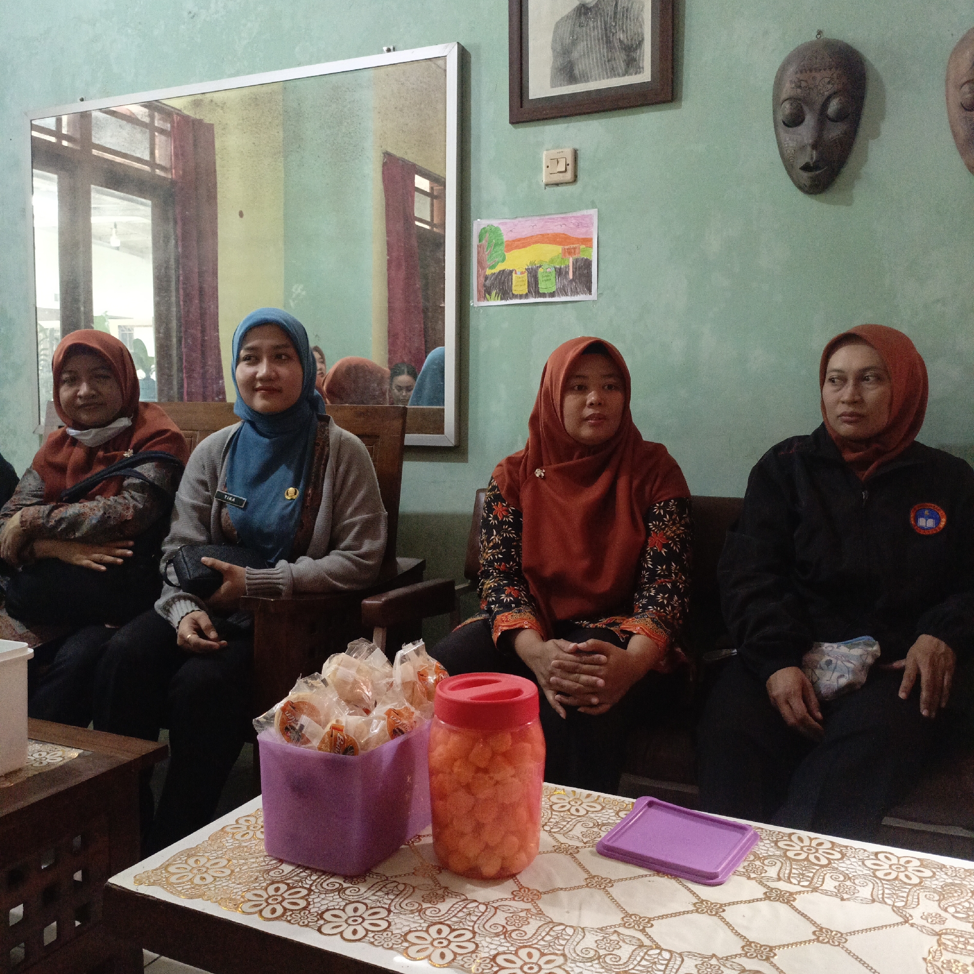 In a dimly lit room with light green walls, four women are seated on wooden chairs. The woman on the far left wears an orange hijab and a patterned shirt. Next to her, a woman in a blue hijab is seated, holding a black clutch bag. She wears a grey cardigan over a dark shirt and has a nametag that reads "TIKA." The third woman from the left, wearing an orange hijab and a black and orange patterned shirt, is seated with her hands clasped. The woman on the far right, in an orange hijab and a black jacket with a circular emblem, is also seated. In the foreground, a table is covered with a lace-patterned tablecloth. On the table sits a purple plastic container filled with packaged snacks, a clear plastic jar with small orange candies, and a small purple tray. A mirror on the back wall reflects a window and part of the room. Artwork and decorative masks are displayed on the walls.
