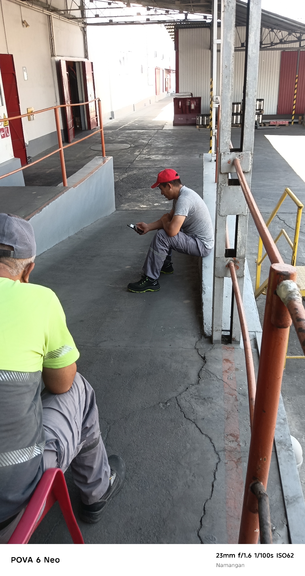 The image shows two men outdoors in an industrial or warehouse area. One man, wearing a neon yellow shirt and a gray cap, is seated on a red stool in the foreground, facing away from the camera. The other man, wearing a gray t-shirt and a red baseball cap, is seated on a concrete ledge in the middle ground, looking down at his phone. He is wearing gray pants and black and green athletic shoes. The ground is asphalt with visible cracks. In the background, there are large red doors, corrugated metal walls, and industrial structures. The overall environment suggests a loading dock or storage facility. The text overlay indicates the location is Namangan City, Uzbekistan.