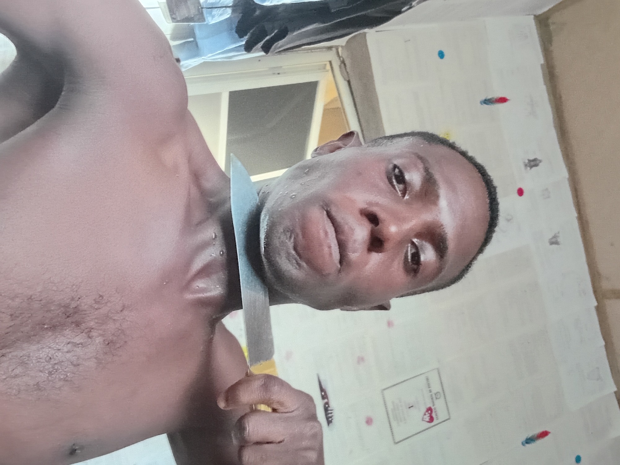 A shirtless man is holding a knife to his throat. He appears sweaty. The background includes a window and a wall covered with papers. There is no indication of this image being taken in Akwete, Nigeria.