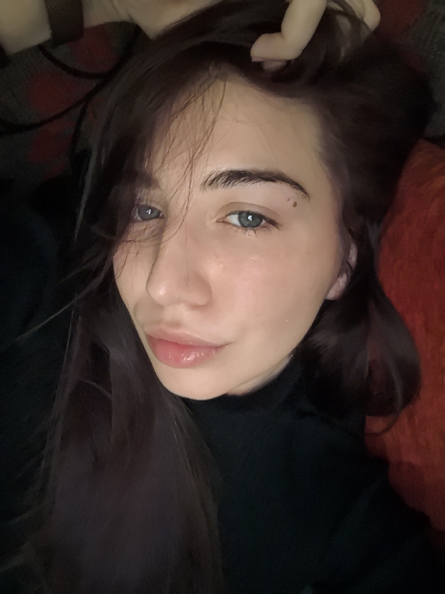A young adult female is depicted in a close-up, slightly elevated shot, lying down indoors. Her head is tilted, and she gazes directly at the camera with light blue eyes. Her dark brown hair, parted slightly to the left, frames her face, with strands falling across her forehead and right cheek. A small, dark mole is visible on her left cheek, below her left eye. Her lips are full, glossy, and slightly parted. She is wearing a dark, high-necked garment. To her right, a soft, textured dark red material is visible. To her left and behind her head, a dark grey fabric with a subtle red and black plaid pattern is present. A portion of her hand or arm, possibly holding her hair, is visible near the top right of the frame. The scene is located indoors in the city of Stara Zagora, Bulgaria.