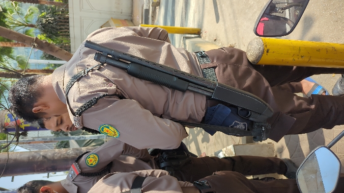 A police officer in a brown uniform with a green and yellow patch stands with a shotgun slung across his back. The firearm is black with a pump-action mechanism. Another officer is partially visible in the background. A motorcycle mirror is prominent in the foreground. A yellow pole is next to a man's brown pants. The setting appears to be outdoors on a sunny day.
