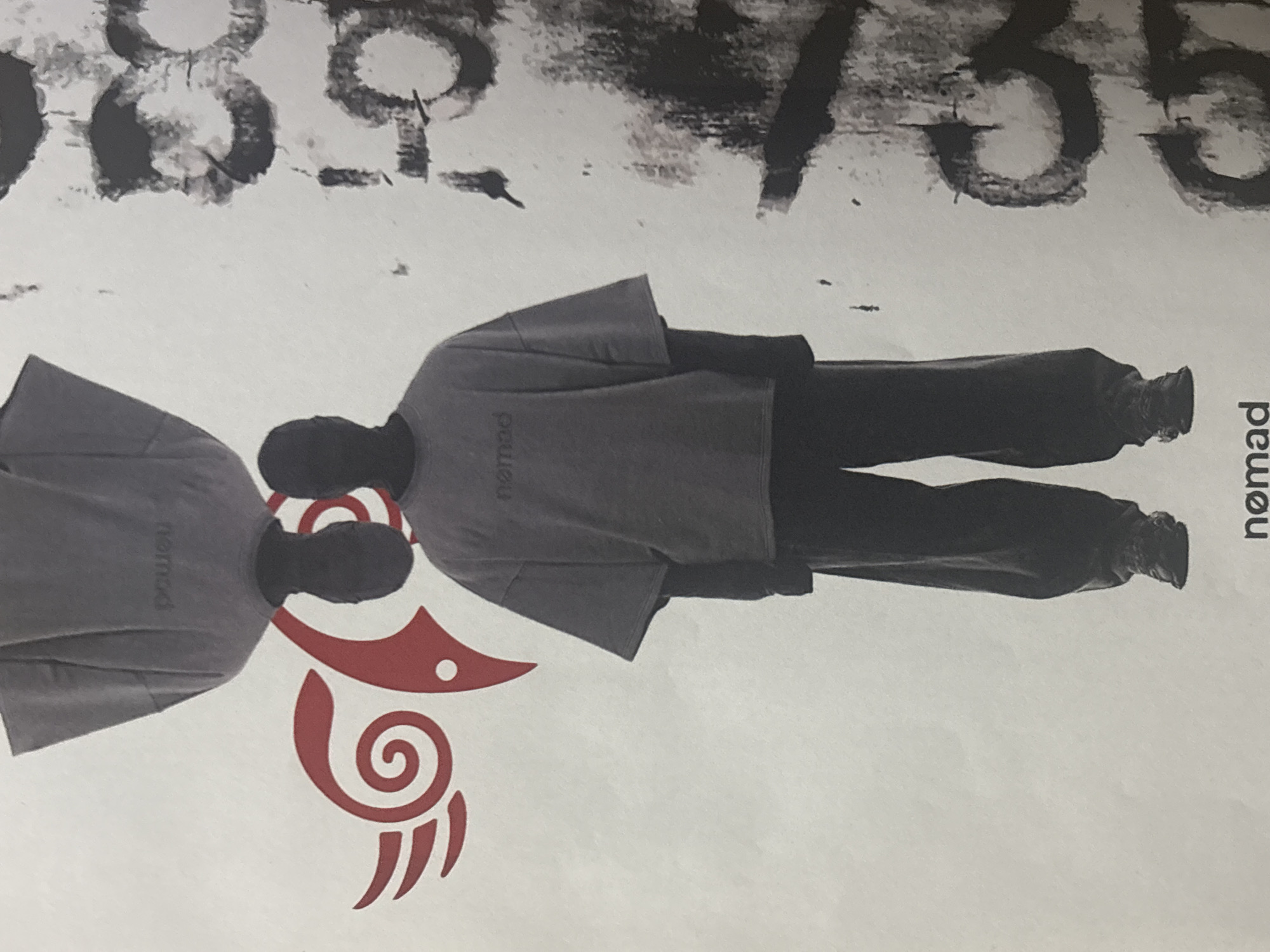 The image displays a minimalist and stylized artwork, likely a poster or print, featuring two human figures and graphic elements against a light background.
**Subjects:** Two human figures are depicted as dark silhouettes wearing oversized, light grey short-sleeved t-shirts and dark, loose-fitting trousers. The word "nomad" is visible in a slightly darker grey on the chest of both t-shirts. The figure on the right stands upright, while the figure on the left appears to be a mirrored reflection, positioned with their heads close together.
**Objects:** A prominent red, stylized graphic is situated centrally between and below the two figures. This symbol features a large spiral, an eye-like shape, and abstract wing or feather-like elements, evoking a tribal or ancient design, possibly representing a bird or mythical creature.
**Scene/Setting:** The background is a plain white or light grey. At the top of the image, large, distressed black numbers and characters are visible, resembling stenciled or spray-painted text. These include "089" on the upper left and a partial "/35" (or "735") on the upper right. Along the far right edge of the image, the word "nomad" is printed vertically in black.
**Activity or event:** The composition suggests an artistic or promotional presentation, showcasing the "nomad" brand and its associated symbolic graphic. The figures are posing rather than engaged in an active event.
**Notable details:** The artwork uses strong contrast and limited colors (black, grey, red, white) to create a striking visual. The distressed typography at the top adds a raw, urban aesthetic. Due to the stylized nature of the figures, no specific emotions or facial expressions are discernible. The location context of Troyan, Bulgaria, is not visually indicated within the image itself.