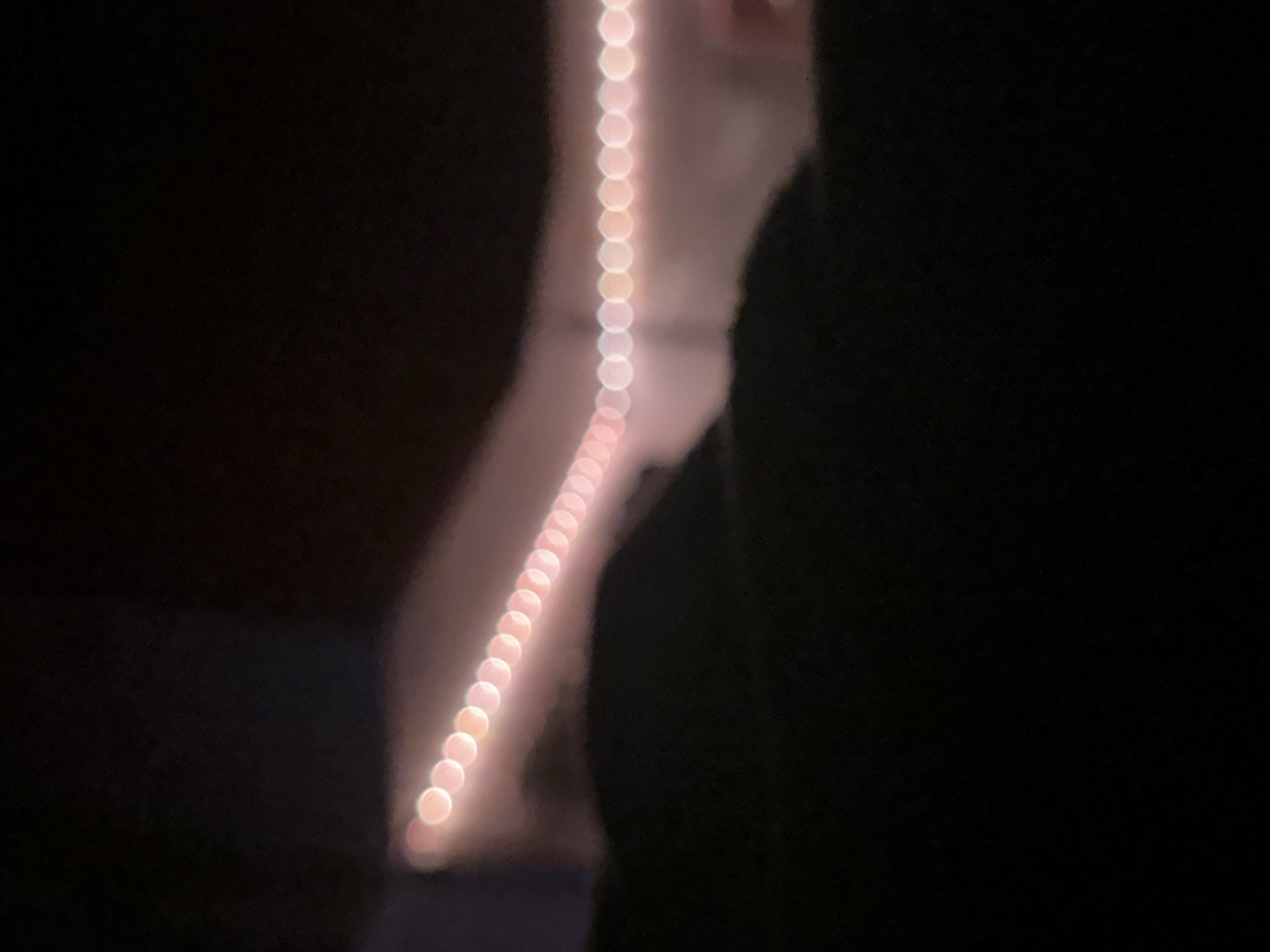 The image depicts a highly blurred and out-of-focus nocturnal scene. A horizontal string of approximately 30 to 35 circular, luminous bokeh lights extends diagonally from the upper left to the upper right. These lights transition in color from reddish-pink on the left to white/pale yellow on the right. Behind the lights, an indistinct, light-toned, blurry structure or surface is visible. The lower two-thirds of the frame is dominated by a dark, amorphous, out-of-focus mass, consistent with foreground foliage or terrain. No individuals, specific objects, or discernible actions are present. Due to the extreme blur and lack of detail, no specific location in Moscow, Russia, or elsewhere, can be identified from the image content.