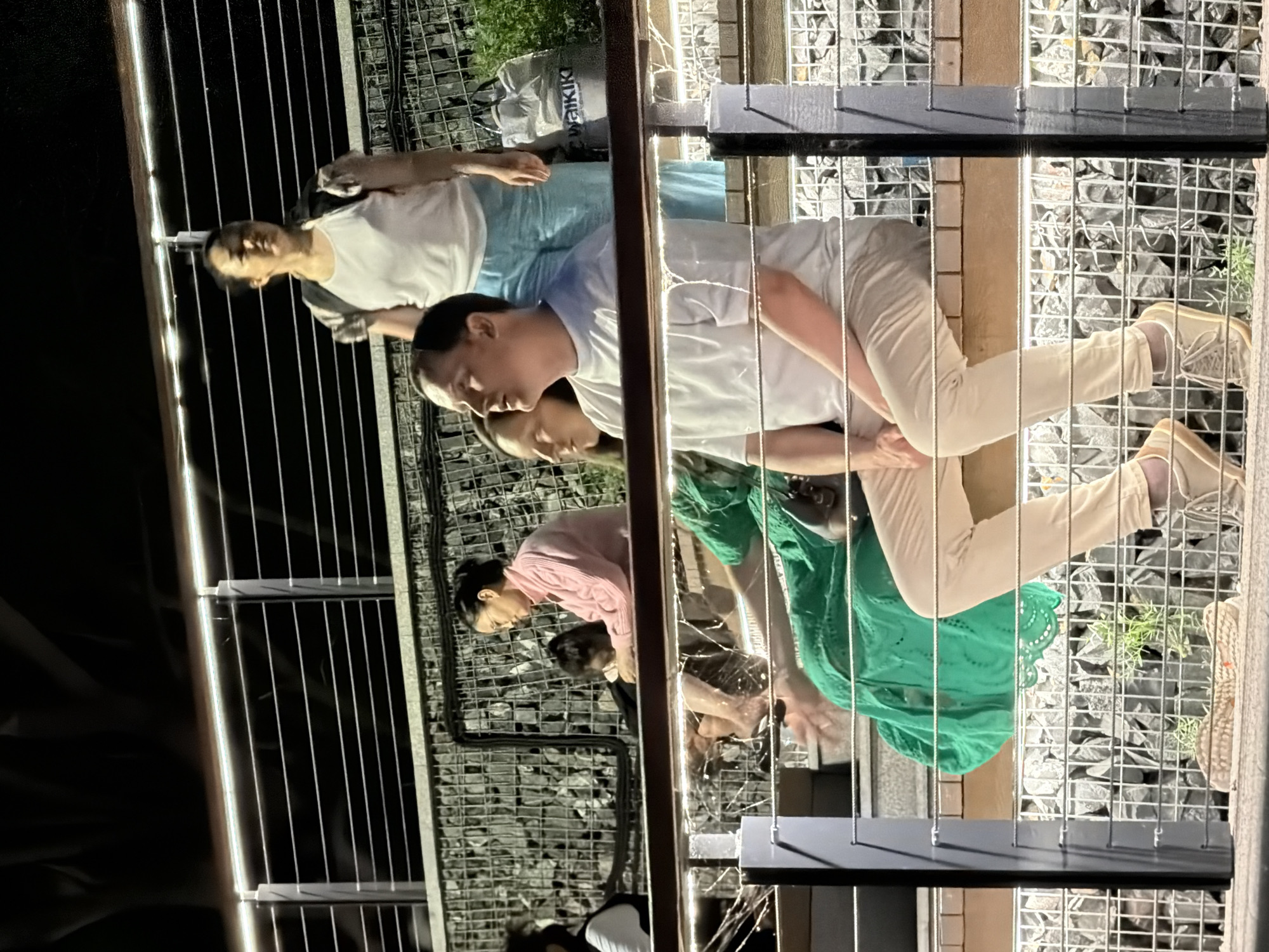 The image depicts a group of people sitting on bleacher-like seating with a railing in the foreground. In the center, a man in a light-colored t-shirt and beige pants is seated, looking to his left. Beside him, a woman in a green dress is also seated, with her arm extended forward. Behind them, another woman in a pink sweater and a child are seated. Further back, a woman wearing a white tank top and blue pants stands with a backpack on. A grey bag with "WAIKIKI" written on it is visible near her. The environment appears to be an outdoor seating area with a stone-filled wall behind the seating. The lighting suggests it is evening or night. The location is identified as the Astana International Financial Centre (AIFC) Green Central Park in Astana, Kazakhstan.