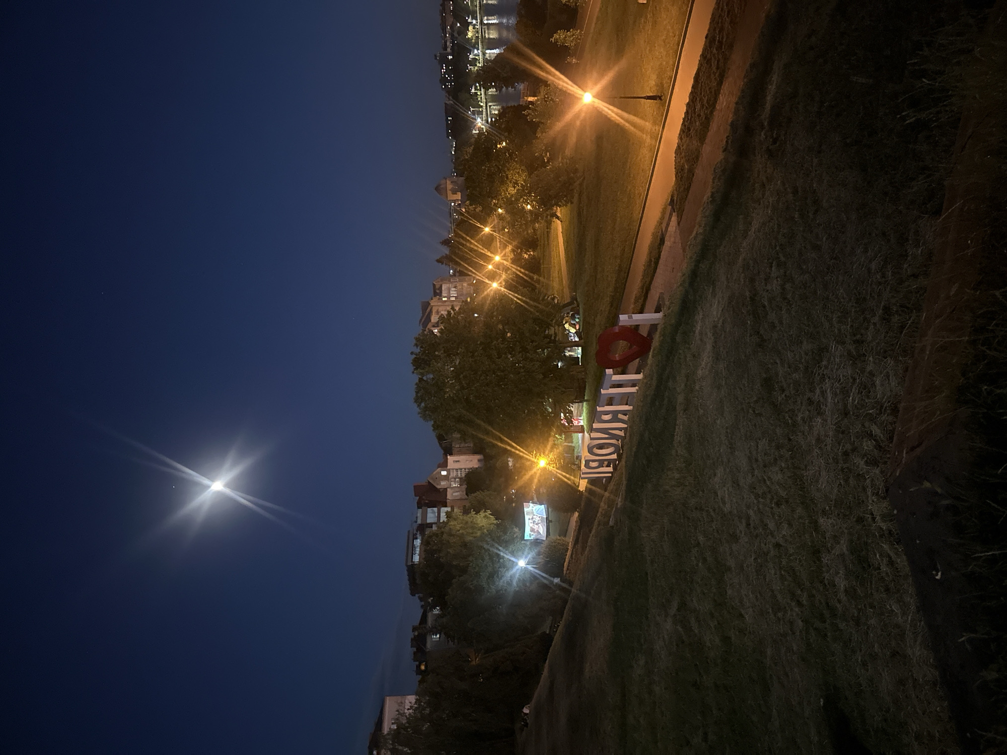 The image captures a nighttime scene in Ternopil, Ukraine. In the foreground, a grassy slope descends towards a paved pathway. To the right of the pathway, large white letters spell out "ТЕРНОПІЛЬ" (Ternopil) with a red heart shape integrated into the design. Beyond the pathway, a park area is illuminated by streetlights, with trees and buildings visible in the background. A large screen is set up in the park, displaying an image. In the far distance, a body of water, likely the Ternopil Pond, can be seen, with lights reflecting on its surface. The night sky is dark blue, with the moon and a bright star or planet visible. The overall atmosphere is calm and serene, with artificial lights providing illumination in the darkness. There are no visible people in the image.