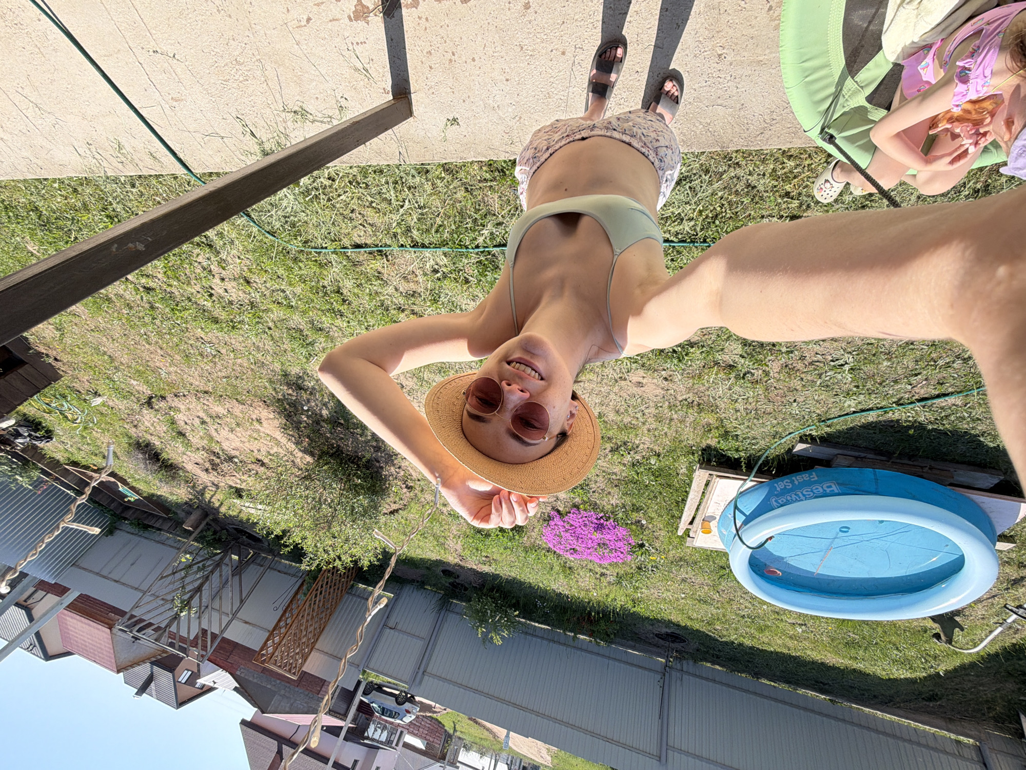A young woman wearing a hat and sunglasses poses for a selfie outdoors. She is wearing a light green top. Behind her is a lawn with a small blue pool and a flower bed. In the background, there are houses and a fence. The location is Chaikovsky, Russia.
