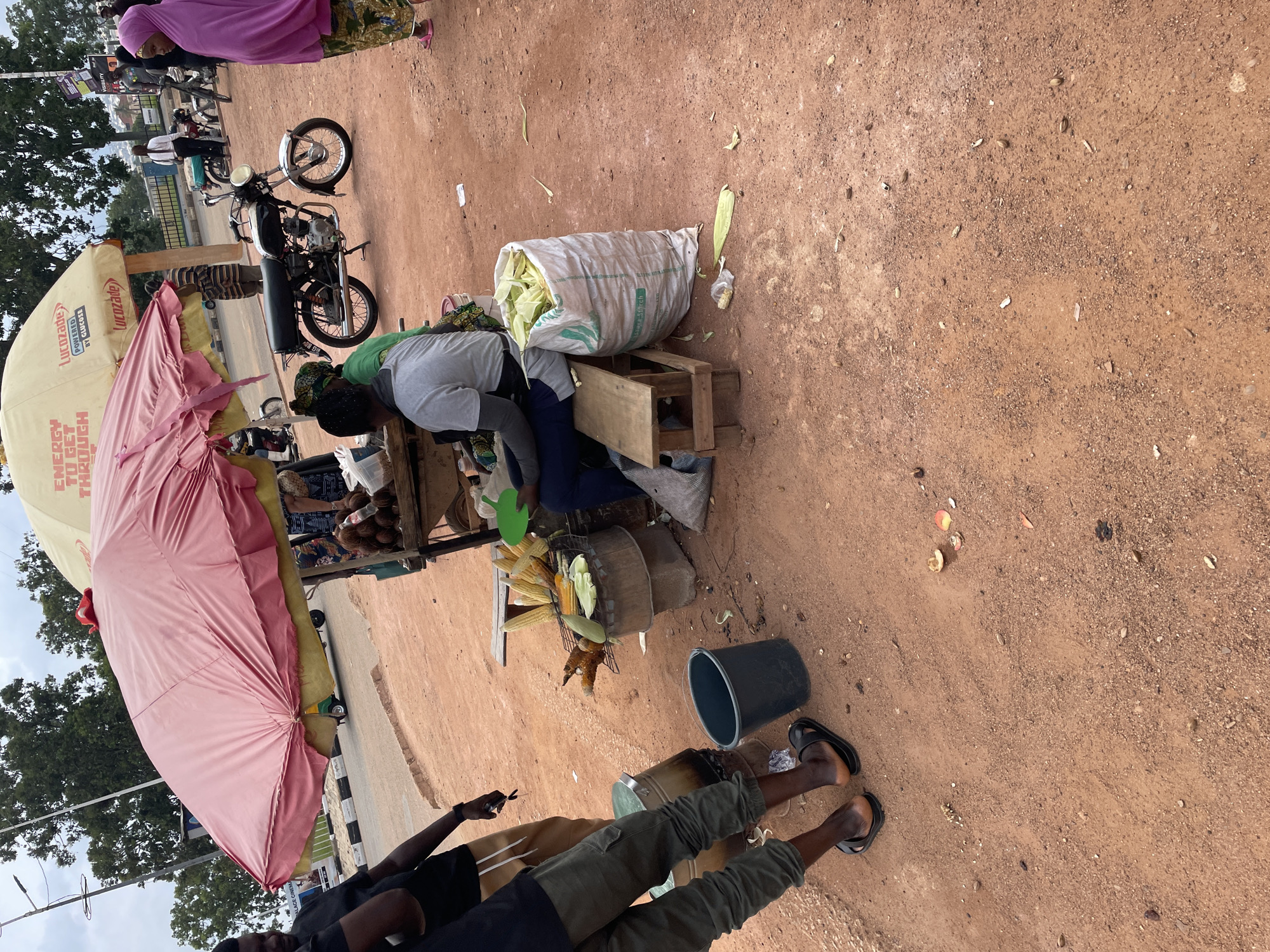 In Bar Jirgi Summa, Nigeria, an outdoor scene depicts a marketplace where corn is being sold and prepared. A prominent pink and yellow umbrella covers a stall, under which several people are gathered. A motorcycle is parked to the right of the stall. The ground is a dusty, reddish-brown dirt, littered with corn husks and other debris. To the left of the stall, two men are standing, one wearing a black t-shirt and the other in a green shirt. A woman in a grey t-shirt is seated behind the stall, attending to the corn. A large white sack filled with corn is placed beside her. In the background, more people and vehicles are visible, suggesting a bustling local market.