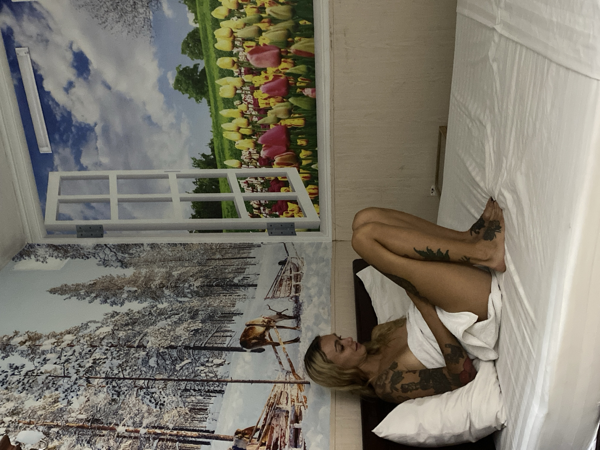 A woman is lying on a bed in a room. The walls are decorated with a mural depicting a winter scene on one side and tulips on the other. The woman has blonde hair and tattoos on her arms and legs. She is partially covered by a white sheet. The bed has white sheets and pillows. The location is Thủ Đức, Vietnam.