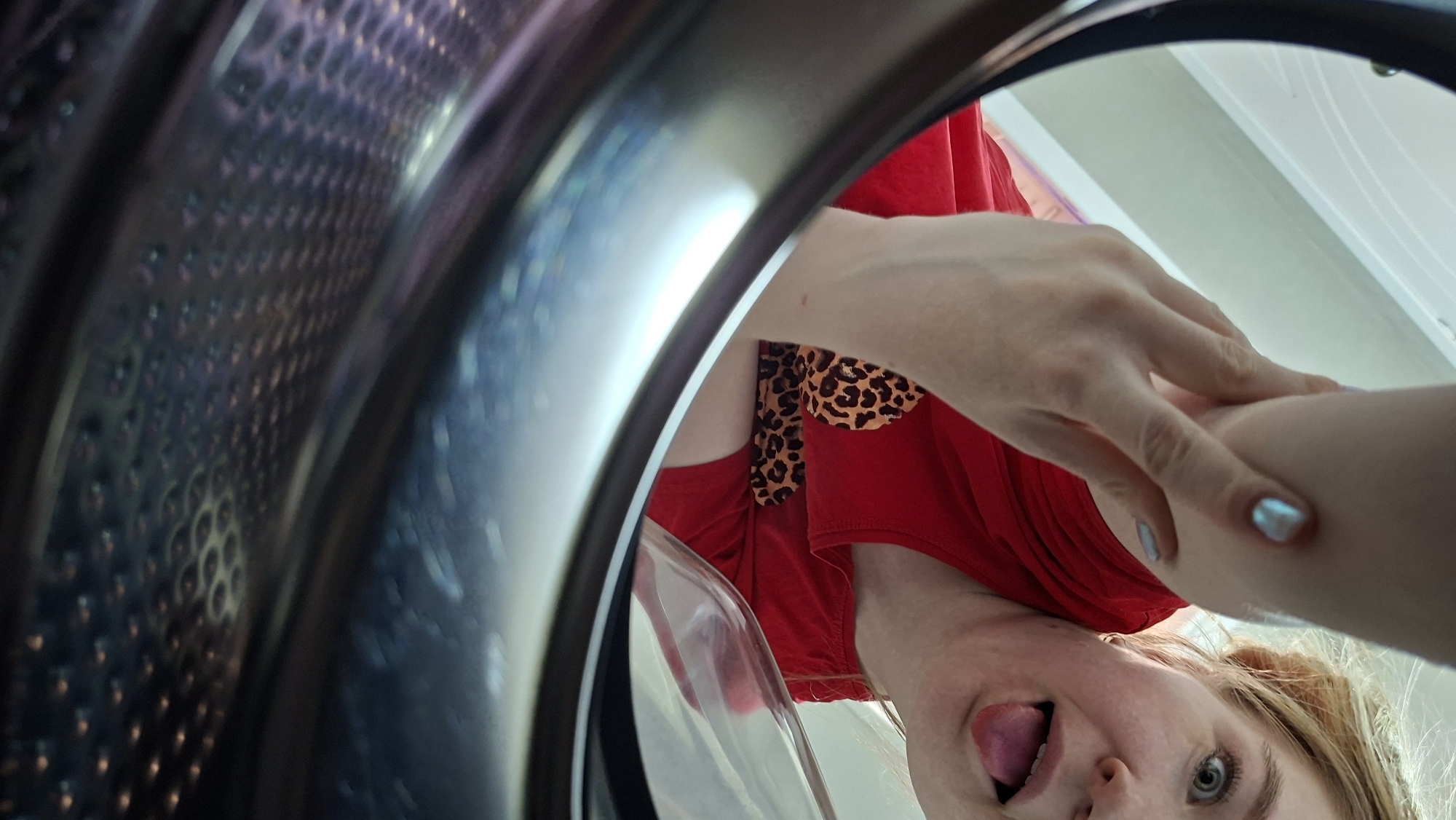 A low-angle close-up shot from inside a cylindrical object, likely a washing machine or dryer. The interior of the cylinder is visible on the left side. A Caucasian woman is looking directly into the cylinder with her tongue sticking out. She is wearing a red shirt with a leopard print accent. Her hands and arms are visible above her head, entering the cylinder. There is a white surface or wall behind her. The location is not identifiable.
