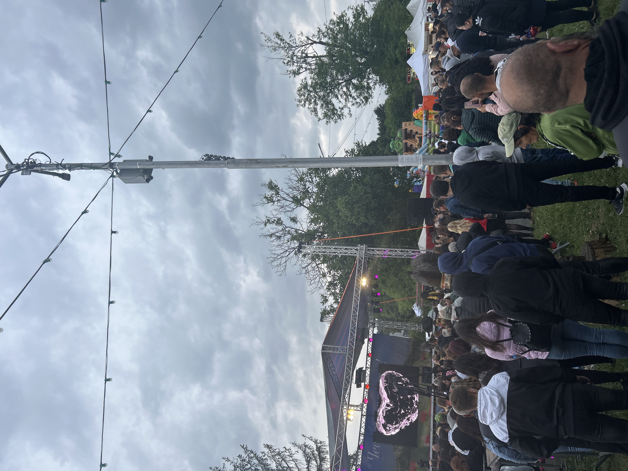 The image depicts an outdoor gathering at a festival or event in Gorna Oryahovitsa, Bulgaria. A large crowd of people is assembled, facing a stage with a large screen displaying a pink heart graphic. String lights are strung across the sky, and a tall utility pole with electrical equipment stands prominently in the center. In the background, trees and tents suggest a park or fairground setting. A bouncy castle with a character's face is visible behind the crowd. The sky is overcast, indicating a cloudy day. The overall scene captures a lively event with many attendees enjoying the festivities.