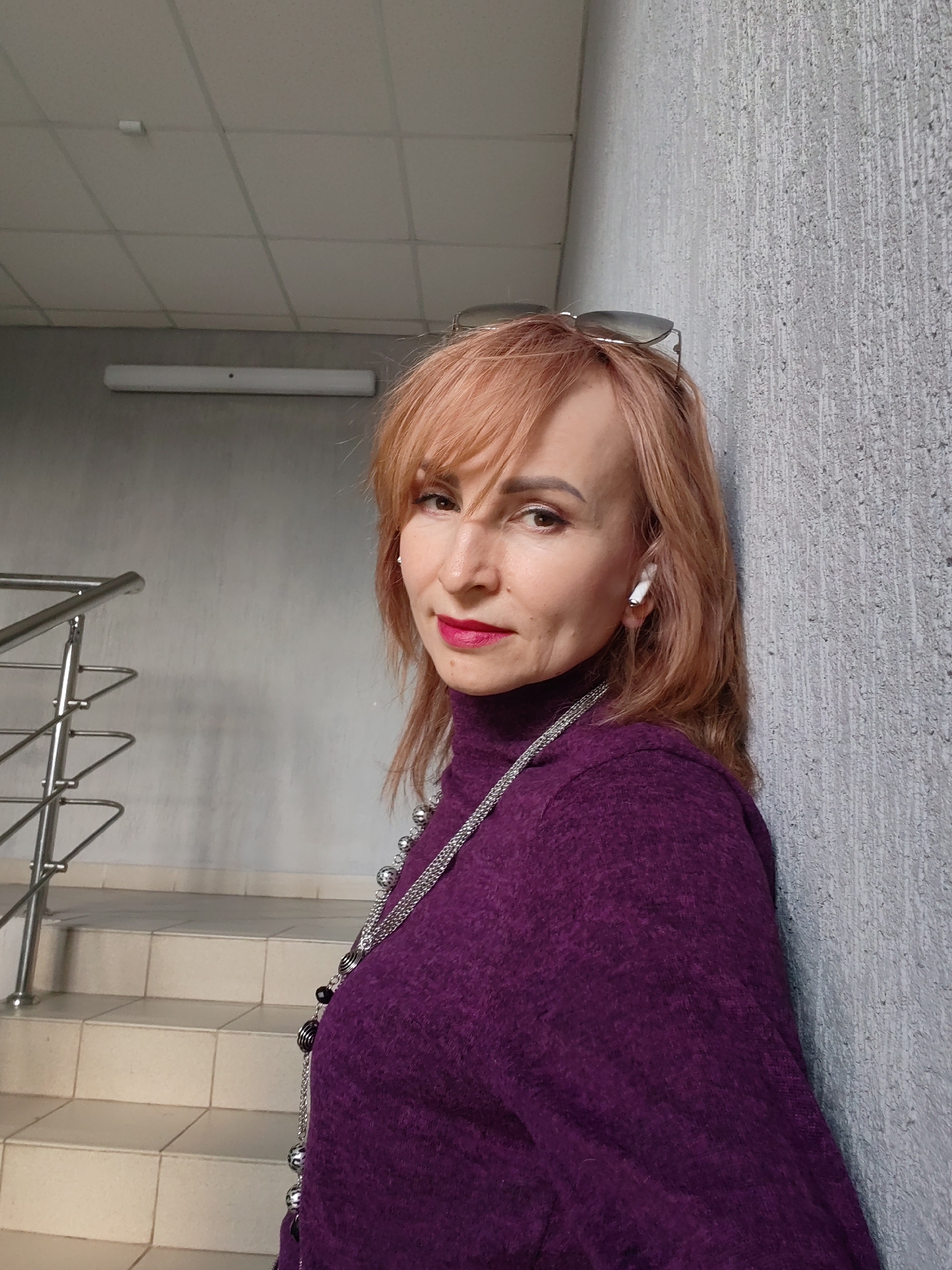 A woman is captured in an indoor portrait, leaning against a textured grey wall. She has light reddish-brown hair with bangs and wears a deep purple turtleneck sweater. A long, multi-strand silver chain necklace with dark, spherical beads is draped around her neck. Sunglasses are perched on top of her head, and a white wireless earbud is visible in her right ear. She is wearing red lipstick and looks directly at the camera with a composed expression.
To the left of the woman, a silver metallic handrail and a light-tiled staircase are visible, suggesting the setting is within a building like an office or public space in Chaikovsky, Russia. The background also includes a smooth grey wall and a white ceiling featuring a grid pattern and a rectangular fluorescent light fixture. The lighting appears to be artificial, common for indoor environments. No text is visible in the image.