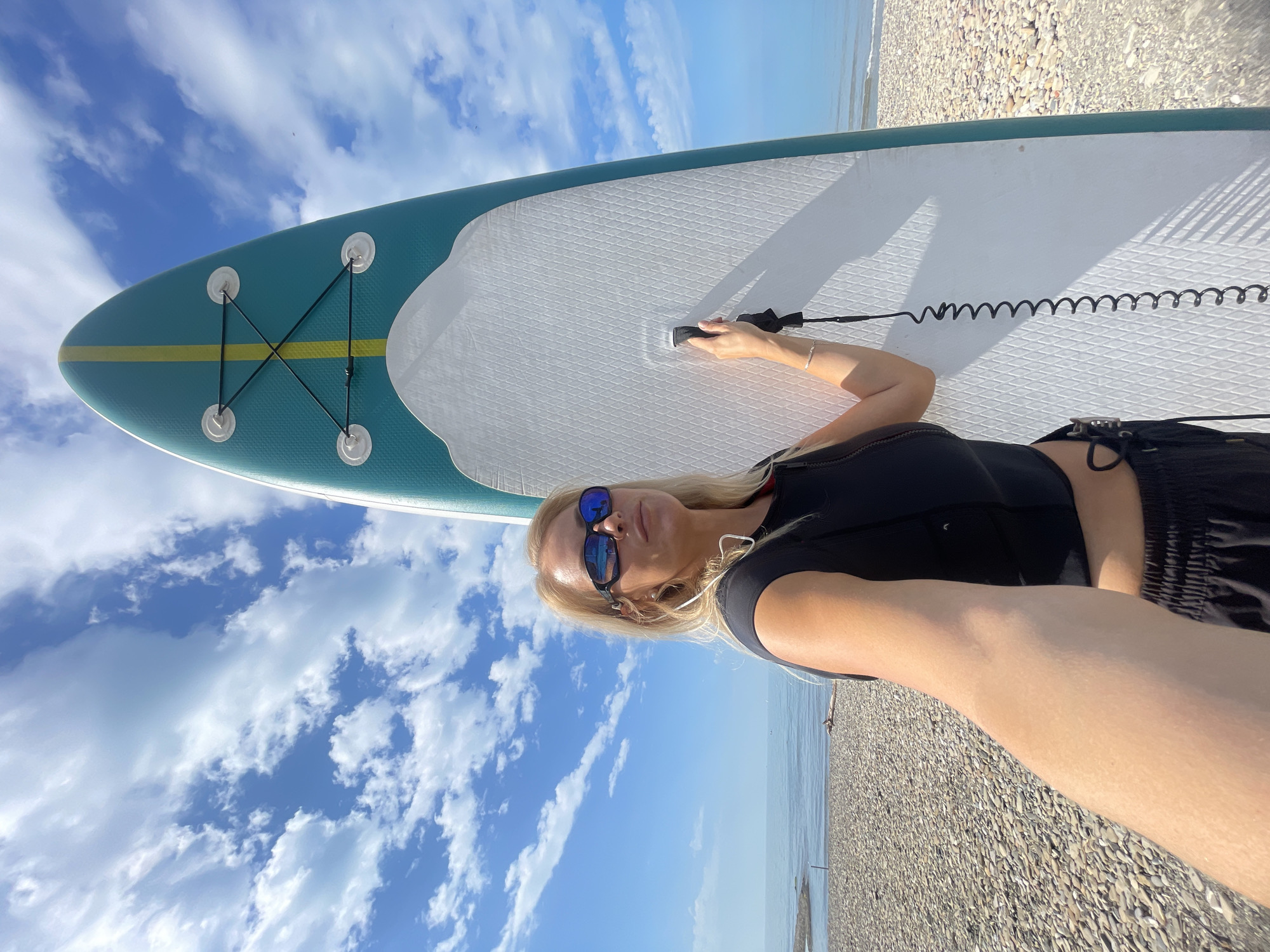 A blonde woman with sunglasses stands on a pebble beach next to a paddleboard. The paddleboard is teal and white with a yellow stripe down the center. The woman is wearing a black crop top and black shorts. She is holding the handle of the paddleboard. The ocean is visible in the background, as is a partly cloudy blue sky. The location is not the city of Прогресс, Russia.