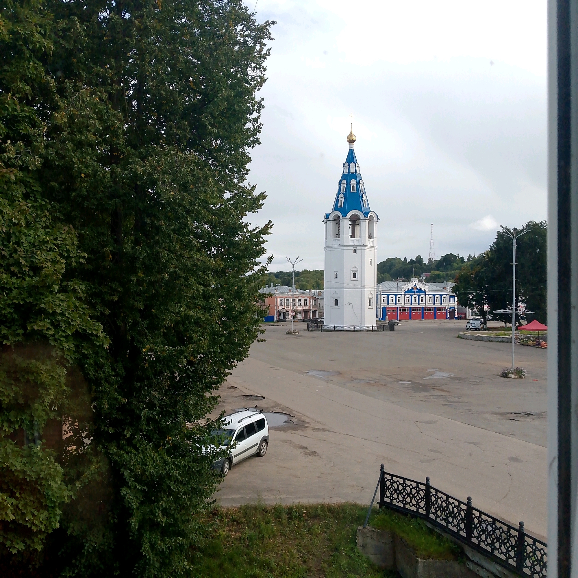 The image depicts a street scene in the city of Vyazniki, Russia, featuring a prominent white bell tower with a blue roof topped with a golden dome. In the foreground to the left, dense green foliage from a large tree partially obscures the view. The bell tower stands in the center of a paved square. Behind the tower, several buildings are visible, including a striking blue and red building with arched doorways, likely a fire station, and other multi-story structures with colorful facades. A white SUV is parked in the square, and a decorative black metal fence runs along the edge of a grassy embankment in the lower right portion of the image. The sky is overcast, suggesting a cloudy day.
