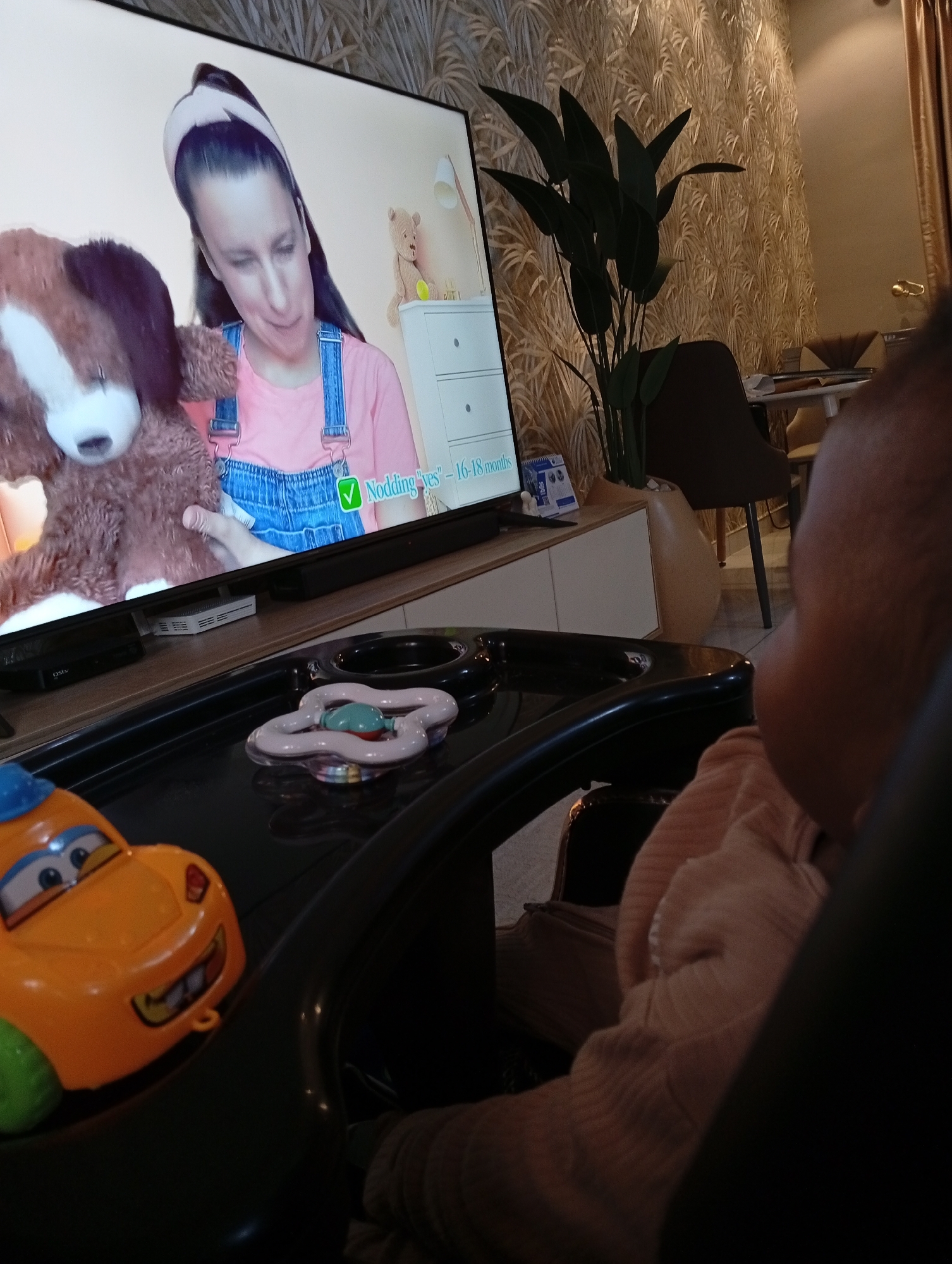 The image depicts an indoor scene with a baby seated in a high chair, facing a television screen. The television displays a woman wearing a pink shirt and denim overalls, holding a stuffed dog. Text on the screen reads "Nodding 'yes' - 16-18 months". In the foreground, a yellow toy car is visible on the high chair tray, along with a colorful baby rattle. The background features a wall with textured wallpaper and a plant. The lighting suggests an ambient indoor environment. The location is stated as Abuja, Nigeria.