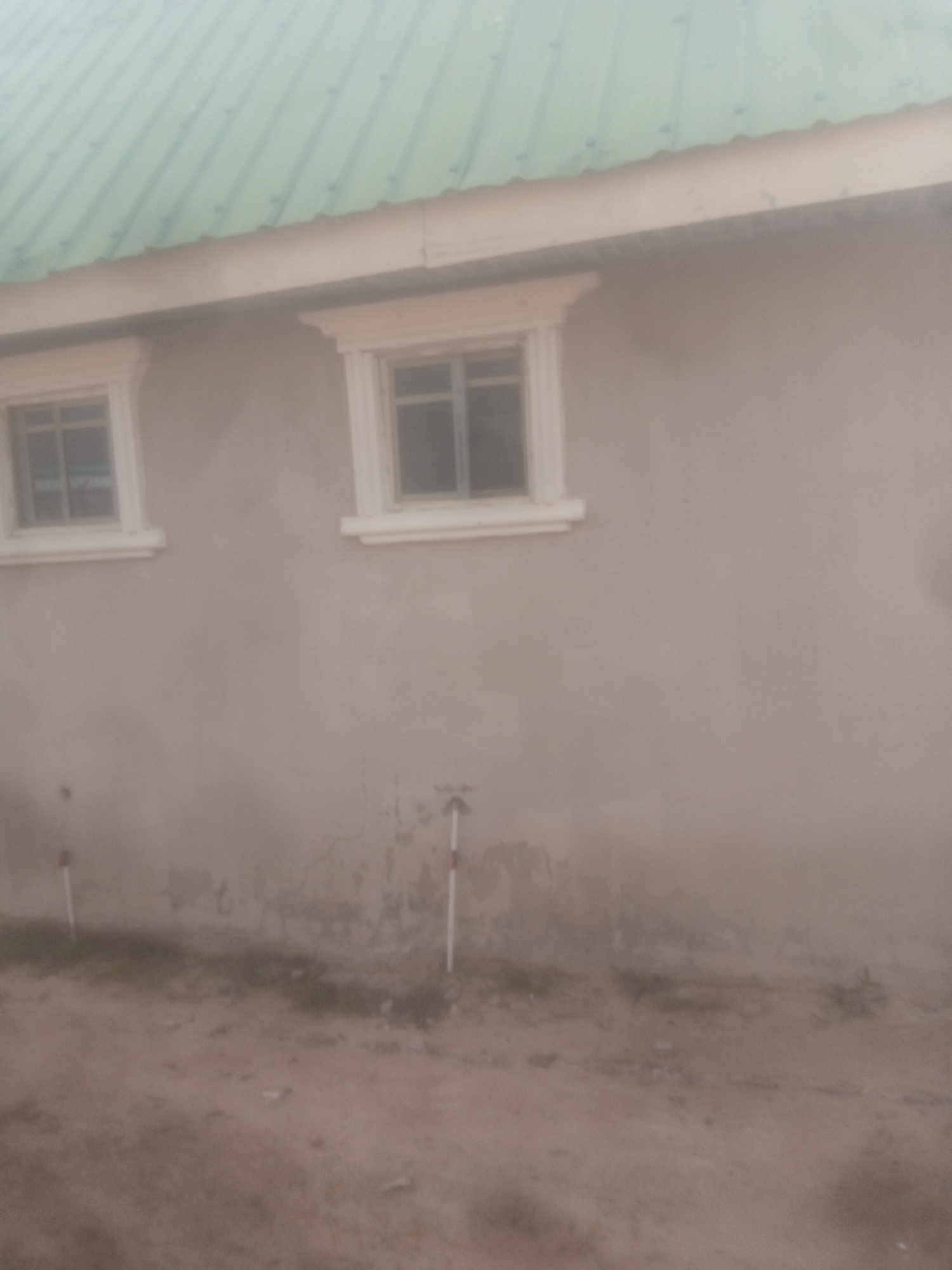 The image depicts the exterior of a building with a green corrugated metal roof and light-colored walls. Two windows with white frames are visible on the wall. The ground in front of the building appears to be dirt and sparse vegetation. Two white poles with red markings are planted in the ground. The scene is likely an outdoor area in Jalingo, Nigeria, based on the provided context. The image is somewhat blurry and lacks clear details about the specific location within the city, or any individuals present.