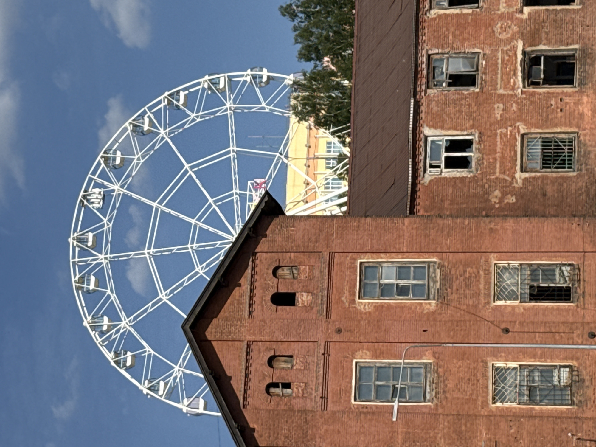 The image shows a white Ferris wheel set against a bright blue sky, partially obscured by the corner of an old brick building. The building is red brick, with visible wear and tear. Some windows are broken, others have bars. It is Izhevsk, Russia.