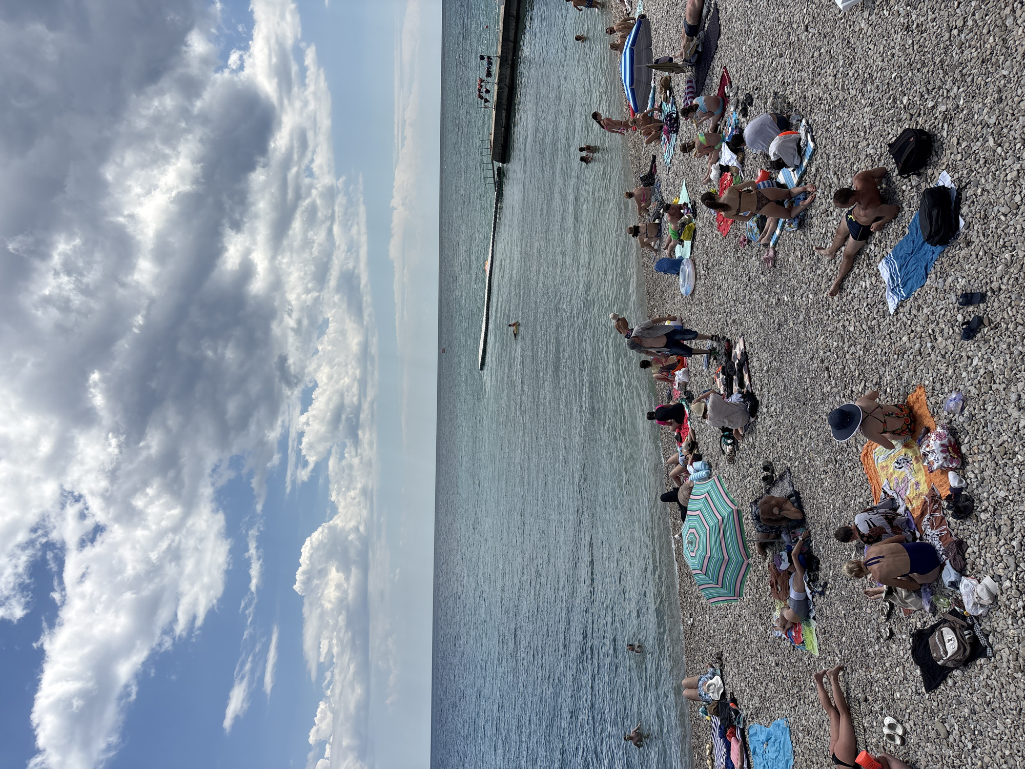 This image depicts a crowded pebble beach in Alushta, Ukraine, under a partly cloudy sky. The water is a calm, deep blue. Numerous individuals are sunbathing on towels and mats, while others are wading or swimming in the sea. A long pier with a platform extends into the water from the right side of the frame, with some people visible near it. Beach umbrellas, bags, and personal belongings are scattered across the beach. The overall scene suggests a typical summer day at a popular tourist destination.