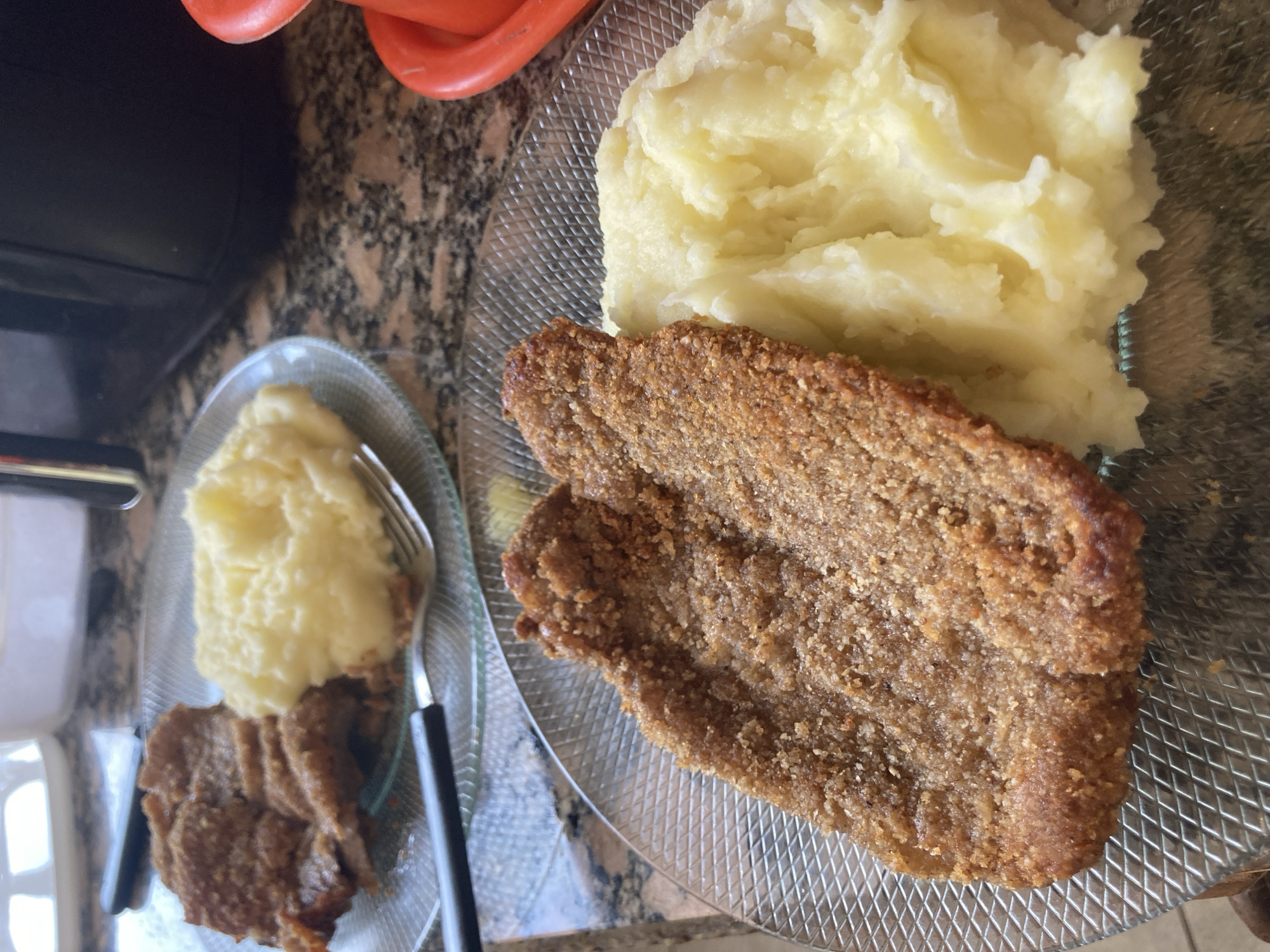 The image shows two plates of food on a patterned countertop. The plate on the left contains mashed potatoes and sliced meat, while the plate on the right contains a larger portion of mashed potatoes and a larger piece of meat. A fork is also visible on the plate on the left. The exact location is unknown.