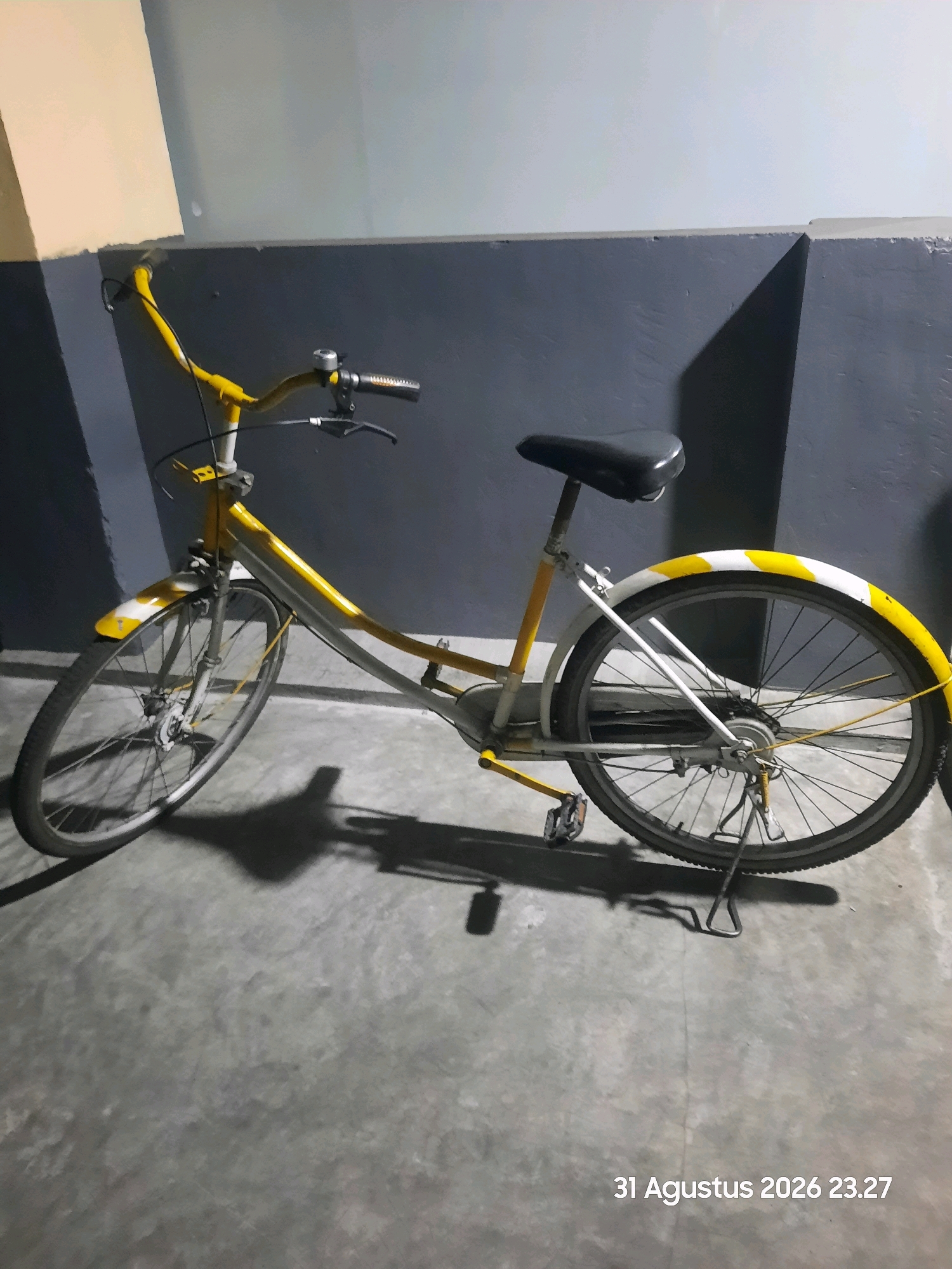 A yellow and white bicycle is parked on a concrete floor in what appears to be an indoor parking area. The bicycle has a dark seat, yellow handlebars, and white fenders with yellow stripes. The background is a dark gray wall with a lighter section at the top. The date and time "31 Agustus 2026 23.27" is visible in the lower right corner of the image, suggesting the location is Gending, Indonesia. There are no people present in the image.