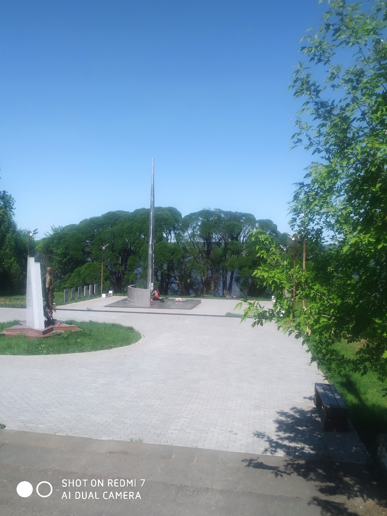 The image captures a view in Ochansk, Russia, featuring a memorial complex. The foreground shows a paved area leading to the memorial. There are two memorials present, one featuring a statue of a figure with a plaque, and another that consists of a tall metal pole with a circular base where wreaths and flowers are laid. Behind the memorial, there is a row of smaller, vertical plaques. A line of trees obscures the view of a body of water in the background. To the right, a tree with vibrant green leaves frames the scene, with a bench sitting beneath its branches. The sky is clear and blue. The timestamp overlay indicates the image was taken with a Redmi 7 AI Dual Camera.