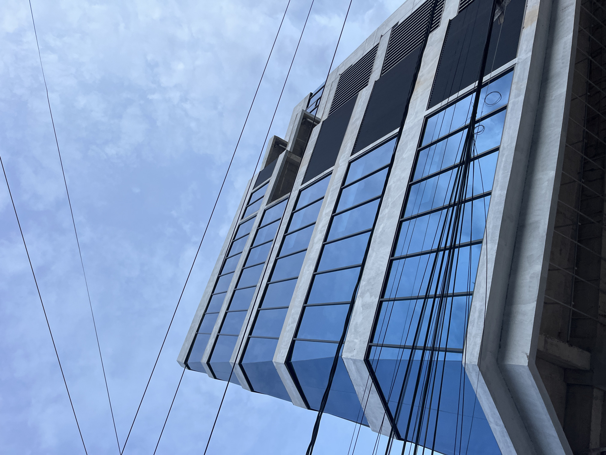 A low-angle shot of a modern building with reflective glass windows in Lhokseumawe, Indonesia. The building's facade is concrete, divided into vertical sections. Power lines crisscross the frame against a cloudy, blue sky.