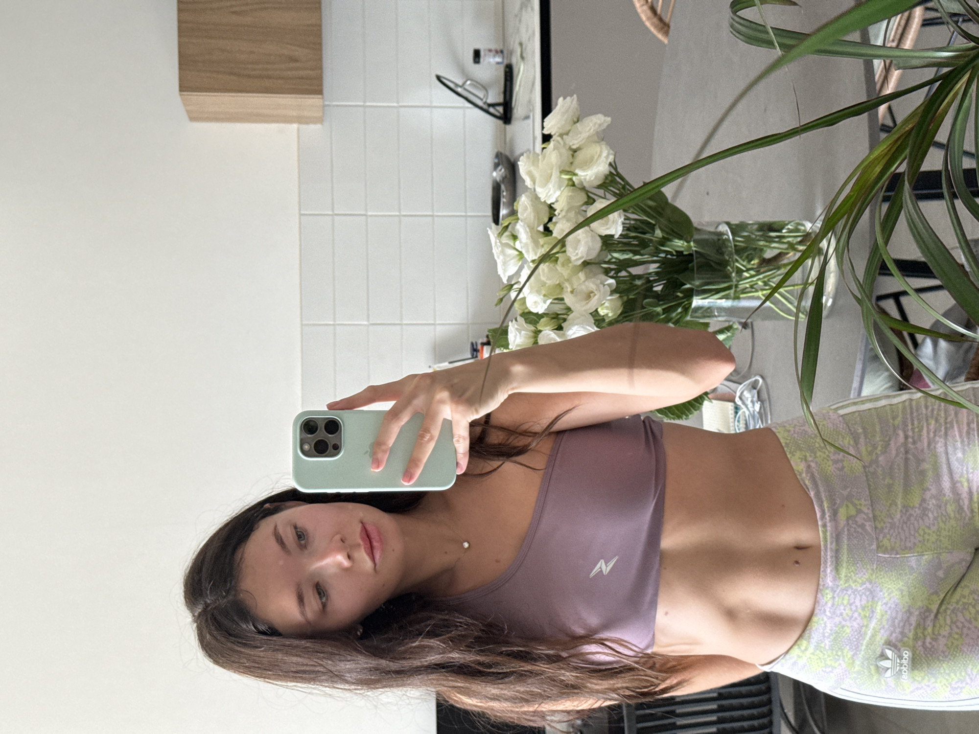 The image, originally rotated 90 degrees counter-clockwise, displays a young woman taking a mirror selfie indoors, likely in an apartment in Tel-Aviv, Israel. She looks directly at the camera with a neutral, relaxed expression, her long, dark brown hair flowing down her shoulders.
She is dressed in a purple sports bra featuring a white, stylized 'N' or lightning bolt logo, paired with light-colored athletic shorts or leggings that have a green and light purple patterned design. An "adidas" logo is visible on her right thigh. Her midriff is exposed, showing a toned abdomen, and she wears a delicate necklace with a small pearl or bead. She holds a light mint green smartphone, equipped with a triple-camera system, in her right hand to capture the reflection.
The background reveals a modern and bright indoor setting, possibly a kitchen or an open-plan living space. Behind her, on a light-toned countertop, stands a large clear glass vase holding a bouquet of white flowers, likely eustoma or roses. A portion of a tall, leafy green houseplant is visible on the right. The wall behind the counter is adorned with white square tiles, serving as a backsplash, while a plain white wall and a wooden cabinet or shelf are seen in the upper left. A section of a wicker chair is also visible on the far right. The scene is well-lit, suggesting daytime.