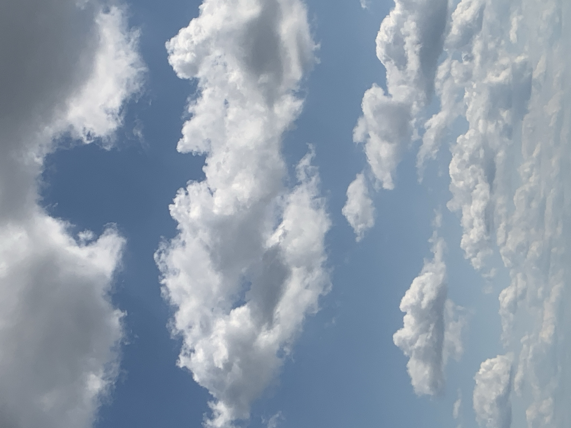 The image is a vertical, medium-angle shot of a partially cloudy sky. The sky is a bright, clear blue, with several distinct cloud formations. The clouds are mostly white and puffy, with some darker gray areas indicating thicker cloud cover or shadows. They are distributed in layers across the frame, with larger, more complex clouds in the upper half and smaller, more scattered clouds towards the bottom. The scene is entirely natural and lacks any man-made structures or human presence. The image does not contain any information that would allow for the identification of a specific location within the city of Kolomyia, Ukraine, or any other geographical area.