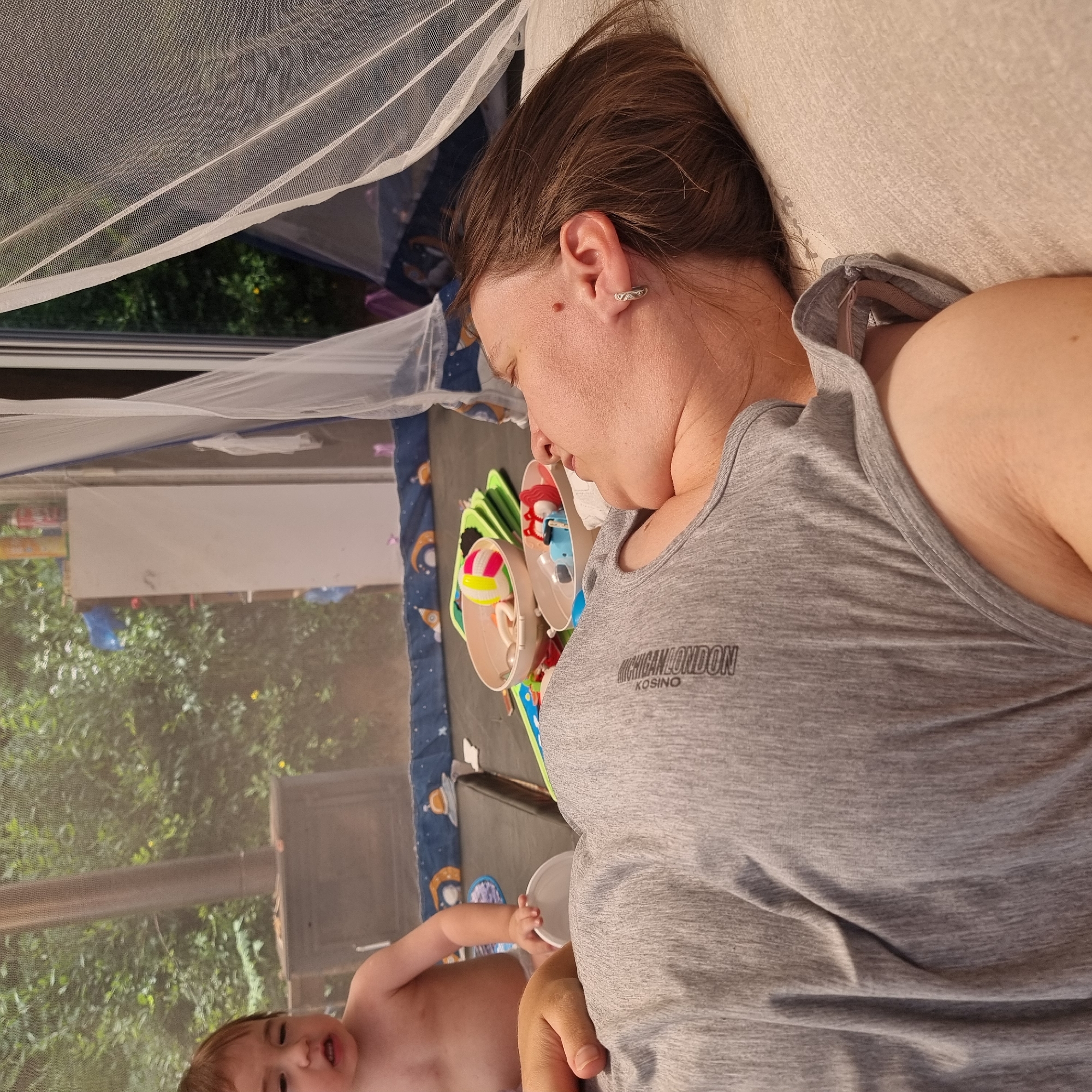 The image shows a woman lying down on her side, facing away from the camera, with her eyes closed. She is wearing a grey t-shirt with a logo that says "CHEF LONDON KOSINO". Beside her, a baby is sitting up and playing with toys. The background is a blurred outdoor scene with greenery and what appears to be a tent or enclosure with a white mesh covering. The overall impression is one of a relaxed, indoor/outdoor domestic setting. The location is not specified in the image.