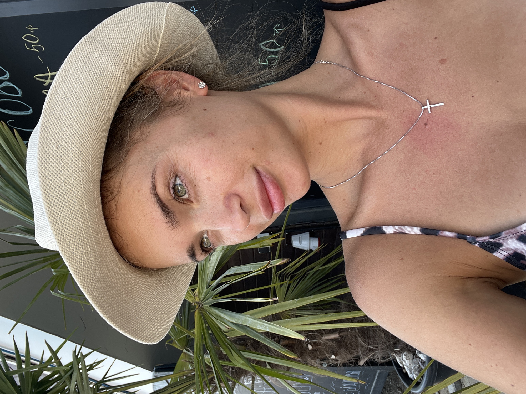 A young woman wearing a wide-brimmed straw hat and a silver cross necklace poses for a selfie outdoors. She has light skin with freckles and green eyes, and her blonde hair is pulled back. She is wearing a black and white patterned bikini top. Behind her, a palm tree with long fronds and a dark chalkboard with writing on it are visible. The lighting suggests it is daytime. The location is identified as Fontanka, Ukraine.
