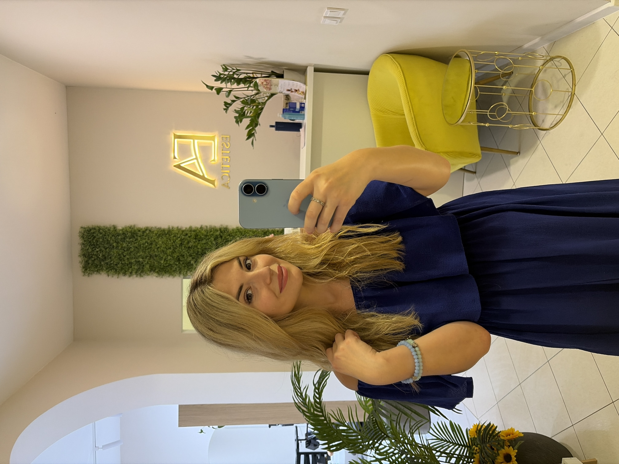A blonde woman with a blue off-the-shoulder dress stands in what appears to be a salon or spa, taking a selfie. She is wearing a bracelet on her left wrist and a ring on her right index finger. Behind her, a bright yellow armchair and a decorative gold side table are visible, along with a potted plant. A neon sign that reads "EA ESTETICA" is mounted on the wall, illuminated in gold. The overall setting suggests a modern and stylish commercial space in Budva, Montenegro.