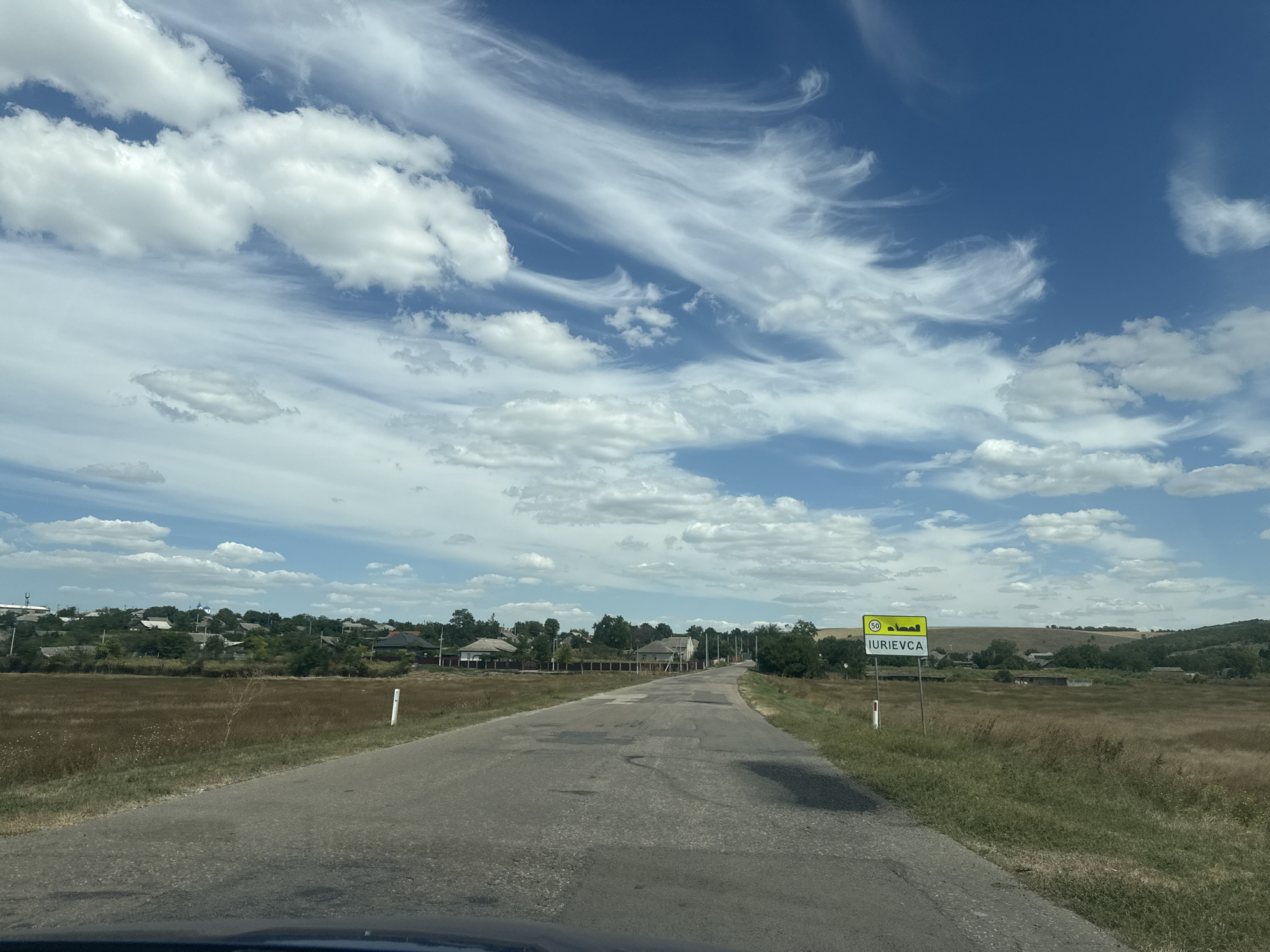 The image shows a rural road leading into the town of Iurievca, Moldova. A speed limit sign of 50 km/h is visible on the right side of the road. The road is paved but shows signs of wear and tear with several potholes. On either side of the road, there are fields of dry grass and scattered trees. In the distance, a cluster of houses with various roof types, along with some greenery and a blue-domed building that could be a church, marks the settlement. The sky is blue with scattered white clouds, some of which are wispy and streaky. The overall scene depicts a sunny day in a quiet, agricultural area.