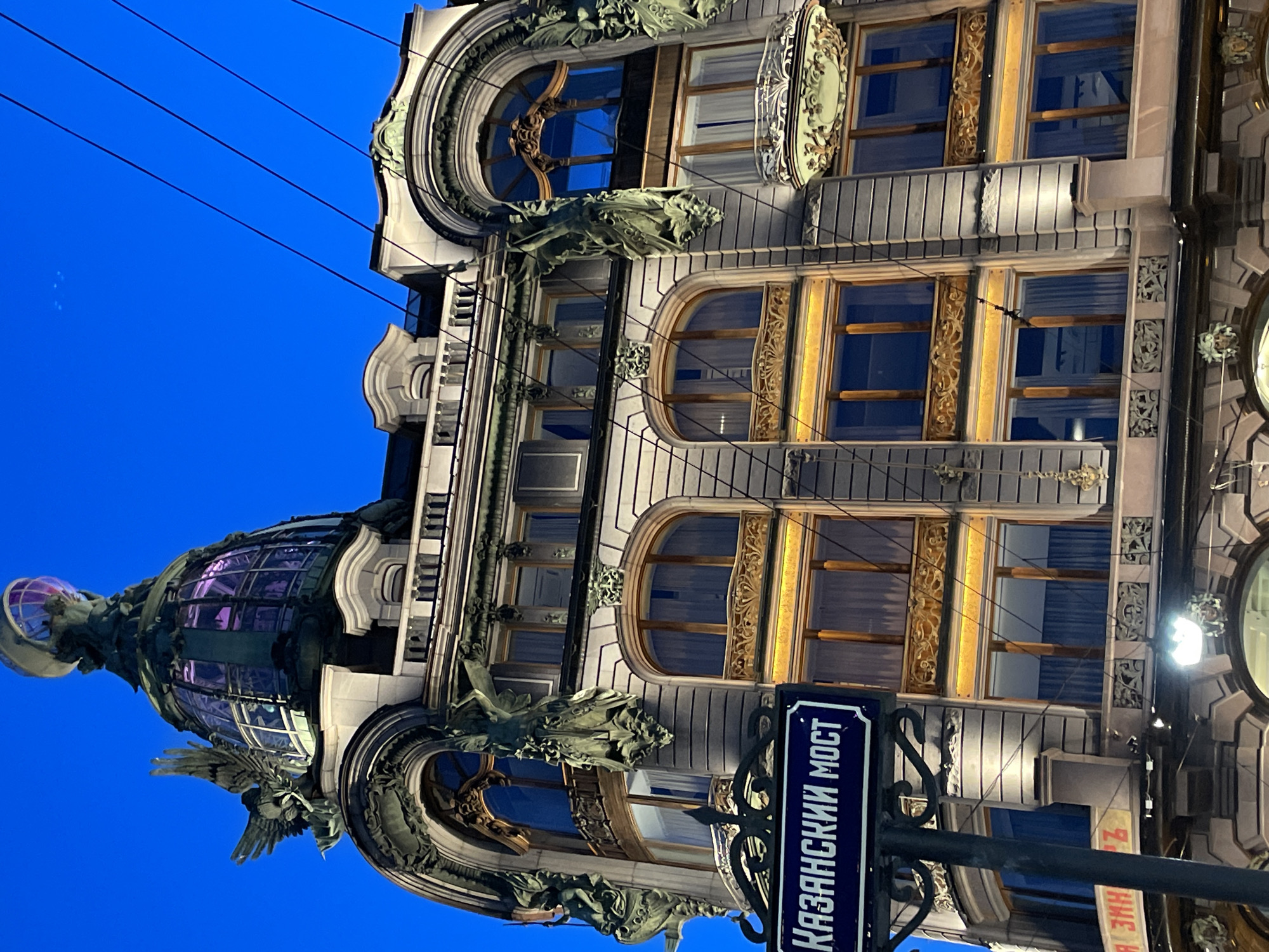 The image displays an ornate, historic building set against a deep, clear blue sky, suggesting dusk or night. The architecture is rich in detail, possibly Art Nouveau, featuring elaborate stone or stucco work, dark sculptural elements, and decorative gold or bronze accents around windows.
The building's facade is adorned with intricate sculptures, including winged creatures that resemble griffins or mythical beasts, positioned beneath windows and at the base of a prominent glass dome. The windows vary in shape, including arched and rectangular designs, many framed with decorative golden elements that catch the light. Interior lights are visible through some windows.
A prominent feature is a large glass dome or cupola on the left side of the building, which emits a soft purple glow from within. This dome is topped with a delicate spire or finial and surrounded by more sculptural figures, possibly angels or eagles.
In the foreground, a dark blue street sign with white Cyrillic text reads "КАЗАНСКИЙ МОСТ" (Kazansky Most / Kazan Bridge), firmly establishing the location as Saint Petersburg. The sign is attached to an ornate black pole. Overhead, several black power lines stretch across the dark sky. The scene emphasizes the architectural grandeur and intricate details of the building, illuminated against the evening sky. No people or specific activities are depicted.