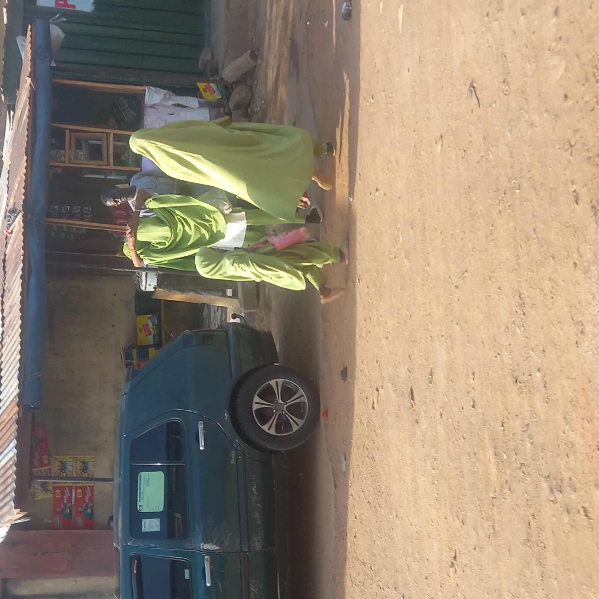 In Dadi Riba, Nigeria, the scene depicts a dark green car parked alongside a building. Two people wearing lime green outfits walk past the car. A building with corrugated metal roofing is next to the car. The ground is dry and dusty.