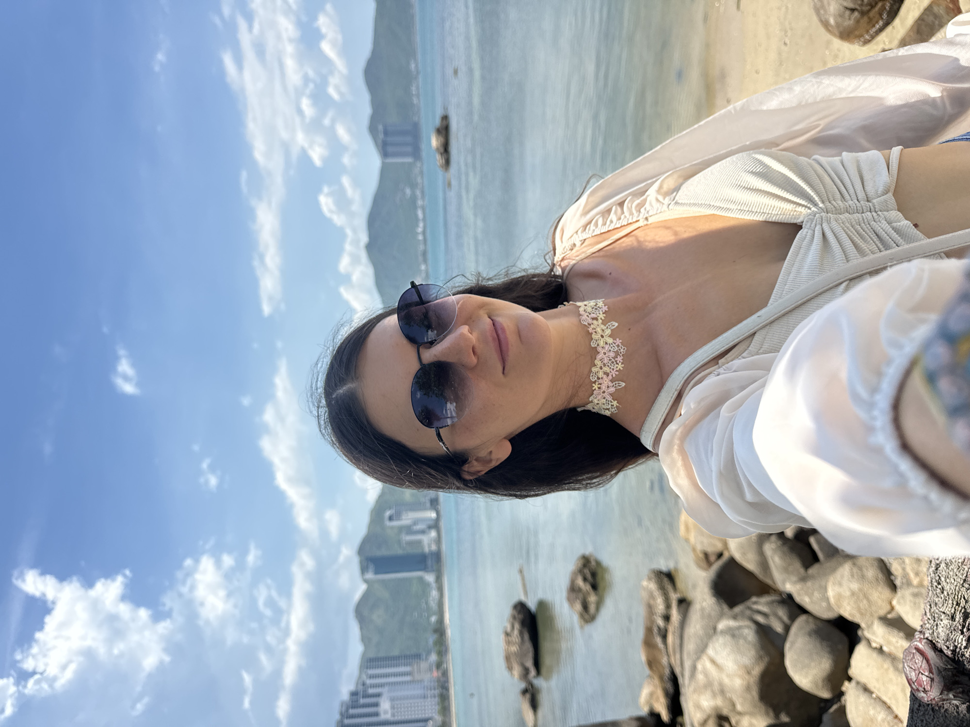 In the city of Nha Trang, Vietnam, a young woman with long dark hair and sunglasses takes a selfie on a rocky beach. She is wearing a light-colored ruched crop top and an oversized sheer shirt. A delicate floral choker adorns her neck. In the background, the calm blue sea stretches towards a distant coastline dotted with tall buildings and lush green mountains under a bright blue sky with scattered white clouds. Large rocks are scattered on the beach and in the shallow water, with some smaller rocks visible in the sea further out.
