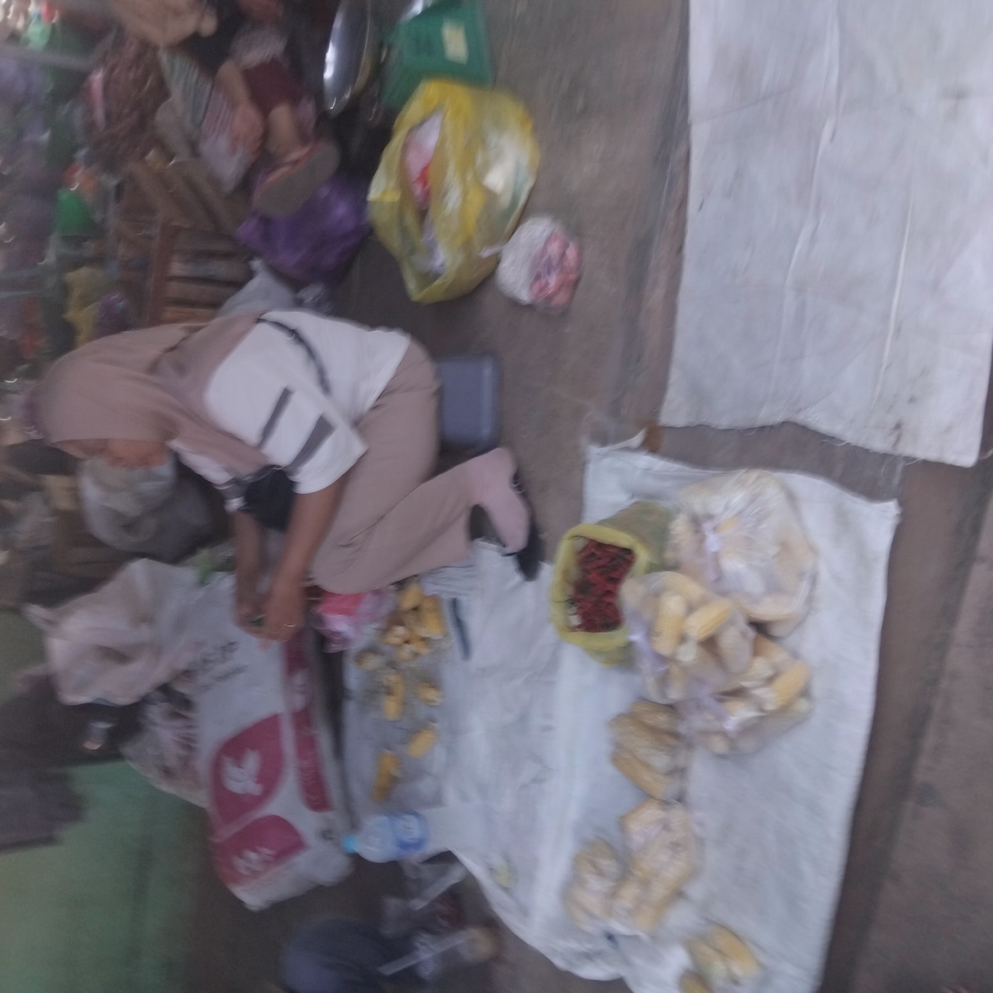 The image depicts a market scene in Pelaihari, Indonesia. A woman wearing a light brown hijab and a white t-shirt with dark stripes is sitting on a low stool. She is facing to the right and appears to be working with her hands, though the activity is unclear due to the image's blurriness. In front of her, on a white cloth spread on the ground, are various items, including packages of what appear to be corn, a bag filled with red chili peppers, and a blue plastic water bottle. Behind her, to the left, is a large white bag with red markings. To the right, a yellow plastic bag is visible, and further to the right, a green weighing scale. The overall environment suggests an outdoor or semi-outdoor market setting with a variety of goods and market equipment.
