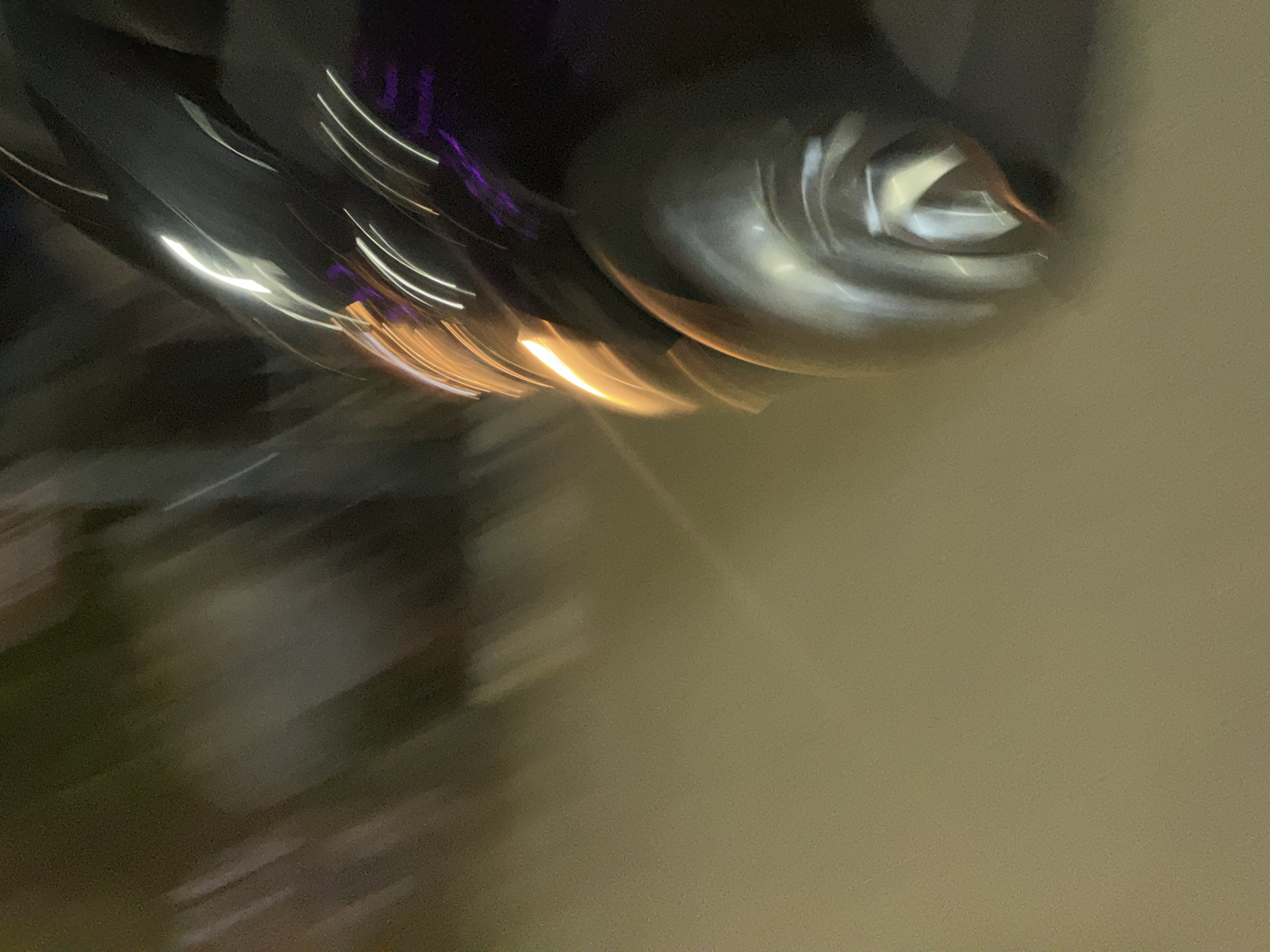 The image is an abstract, motion-blurred photograph of the front section of a dark-colored vehicle, possibly a car, at night. The vehicle's headlight emits a bright orange glow, and streaks of white and purple light from other sources, likely streetlights or other vehicles, traverse the scene. The ground appears to be a muted, light brown or beige surface, possibly asphalt or concrete, reflecting some of the light. The overall impression is one of movement and low light conditions. The image does not provide specific visual cues that would allow for precise localization within Abuja, Nigeria.