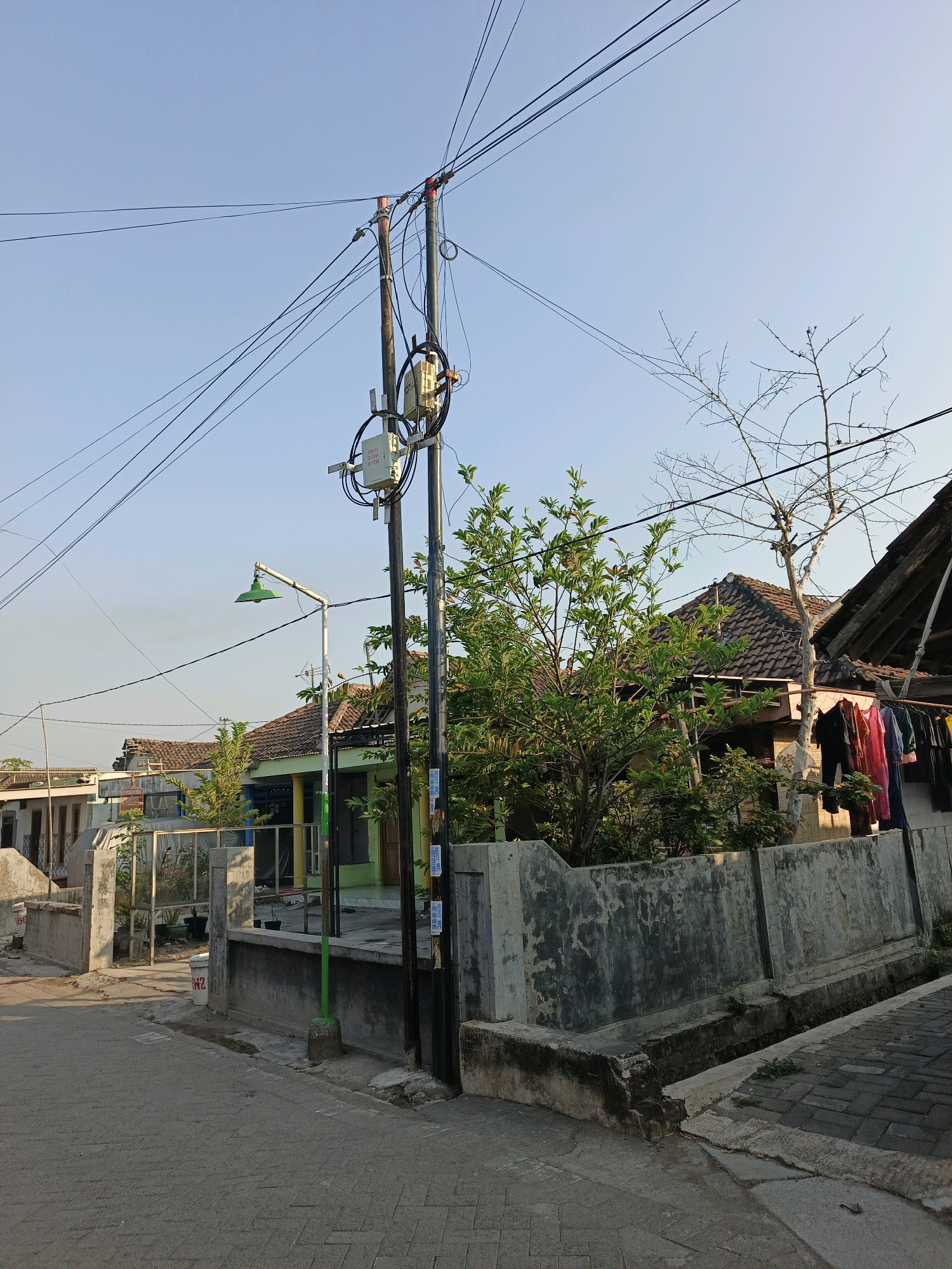 The image depicts a street scene in Ketegan, Indonesia, bathed in the warm light of late afternoon or early morning. The sky is clear and a pale blue. A concrete sidewalk paved with interlocking bricks runs along the right side of the image, adjacent to a concrete drainage ditch. To the left of the ditch, a low, weathered concrete wall runs parallel to the street.
Dominating the center of the frame is a utility pole, dark and covered in a dense network of overhead wires. Attached to the pole are several telecommunication boxes and what appears to be a street lamp with a green conical shade, mounted on a white pole. To the left of the utility pole, a light-colored fence encloses a small area with some potted plants visible.
Behind the fence and wall, several houses with traditional tiled roofs are visible. One house, painted green, has a colorful awning or overhang. To the far right, a section of a house features an open roof or patio area, with laundry hanging on a clothesline, including red, blue, and black garments. A bare, spindly tree stands next to this area, its branches stark against the sky. Lush green foliage from a leafy tree is prominent in the foreground and midground, obscuring parts of the houses and creating a contrast with the bare tree. The street itself is paved with interlocking bricks, and a gray concrete wall with visible signs of wear and tear borders the immediate foreground.