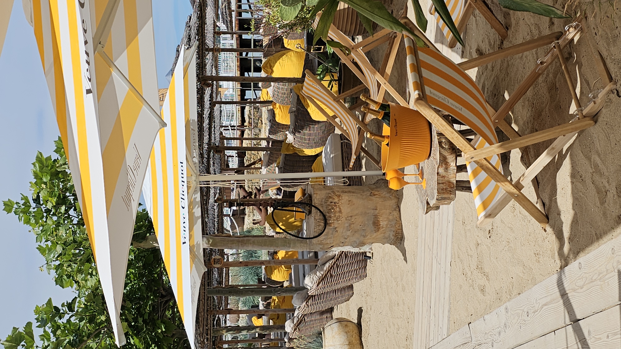 This media file depicts a sunny beachside setting, likely a restaurant or bar area. The scene is characterized by its bright, summery atmosphere.
Prominently featured are several yellow and white striped beach umbrellas and deck chairs, creating a cohesive and festive visual theme. One umbrella has the text "Veuve Clicquot" visible, indicating a possible sponsorship or branding. In the foreground, a bright orange Veuve Clicquot bucket holds bottles of champagne, accompanied by two matching champagne flutes, suggesting a celebration or a luxurious experience.
In the background, there are shaded seating areas with thatched roofs, furnished with woven chairs and cushions, some of which are yellow. Wooden structures and natural materials like sand and wood planks contribute to the rustic yet chic beach ambiance. Green foliage from trees or plants is visible, adding a touch of nature to the scene.
The image appears to be taken during the daytime, under clear, sunny skies, as evidenced by the strong shadows and bright lighting. The overall mood is relaxed and inviting, typical of a beach vacation spot. While no people are clearly visible and actively engaged in an activity, the setup strongly implies a place for leisure and enjoyment. The location context provided is Varna, Bulgaria, which aligns with the beach setting.