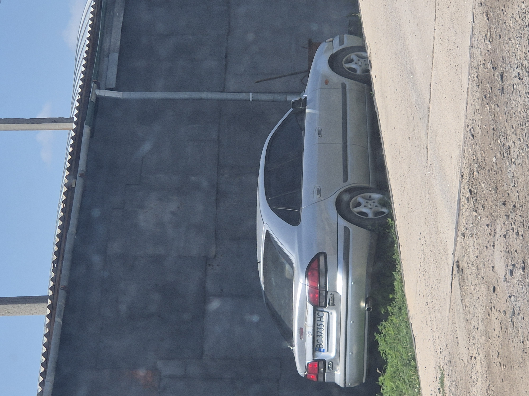A silver Mazda 626 sedan is parked on the side of a dirt road, partially obscured by a gray concrete wall. The car's license plate, visible from the rear, reads "BC 3735 HC". A corrugated metal roof overhangs the wall, with a drainpipe running down its side. Green vegetation grows along the edge of the road and behind the car. The scene appears to be in an outdoor, possibly industrial or rural, area in Pidhaitsi, Ukraine. No people are visible in the image.