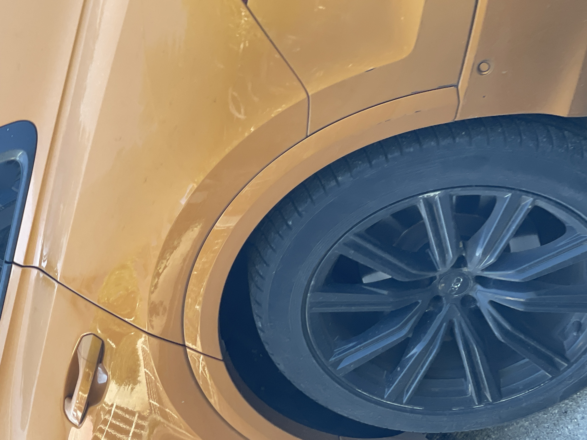 An eye-level close-up shot captures the side-rear section of a golden-yellow or mustard-colored car, parked outdoors in daylight. The image is oriented vertically, showing the car's passenger side with the rear wheel prominently featured.
The car's body panels are a glossy, metallic golden-yellow, exhibiting reflections of what appear to be nearby structures or buildings. The surface shows some dust, smudges, and minor marks, particularly around the fender arch. A distinct dark seam runs horizontally along the car's side, separating different body panels.
The large, multi-spoke wheel is dark, possibly black or dark grey, and fits snugly within the car's robust wheel arch. The tire is black with a visible tread pattern. At the center of the wheel, the four interlocking rings of the **Audi** logo are clearly visible on the hub cap.
In the lower left portion of the image, a car door handle is present, also in the golden-yellow color of the car. Above the wheel well, on the rear fender, a small, circular sensor (likely a parking sensor) is visible. The bright lighting suggests a clear, sunny day. No other specific location cues or activities are discernible.