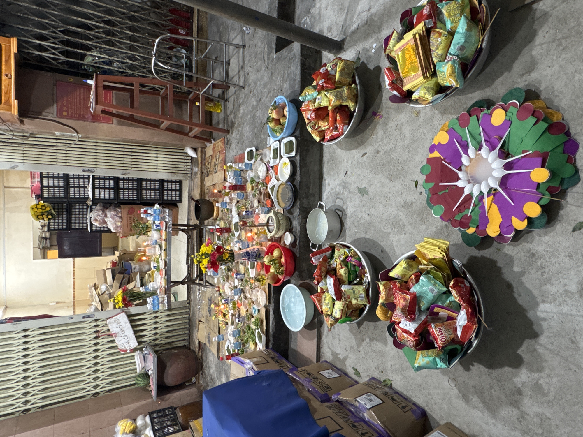 The image depicts an outdoor offering table set up in front of a building with a large metal grate door. The table is laden with various items, including food, drinks, candles, and what appear to be offerings for a religious or cultural ceremony. Several bowls filled with snacks and fruits are prominently displayed on the ground in front of the table. In the background, there are stacks of boxes and a ladder, suggesting a temporary setup. The lighting indicates it might be evening or a dimly lit area. The scene appears to be located in a street or alleyway in Nha Trang, Vietnam.