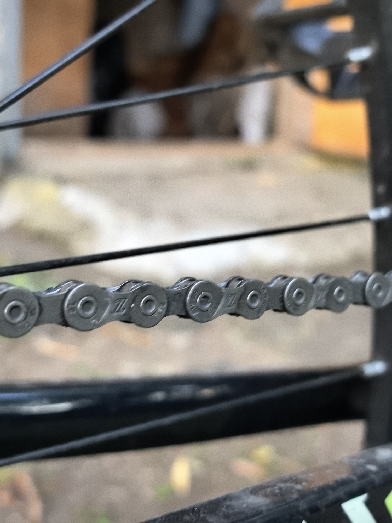 An extreme close-up, eye-level shot focuses on a bicycle chain. The chain is a metallic gray and appears to be made of interconnected links, each with a circular indentation. Some of the links have a faint "Z" logo visible. The chain is horizontal, stretching across the frame, and is supported by thin, dark spokes of a bicycle wheel. The background is blurred and out of focus, showing hints of earthy tones and possibly some greenery, suggesting an outdoor environment. The lighting appears to be natural daylight. There are no people or specific city landmarks visible.