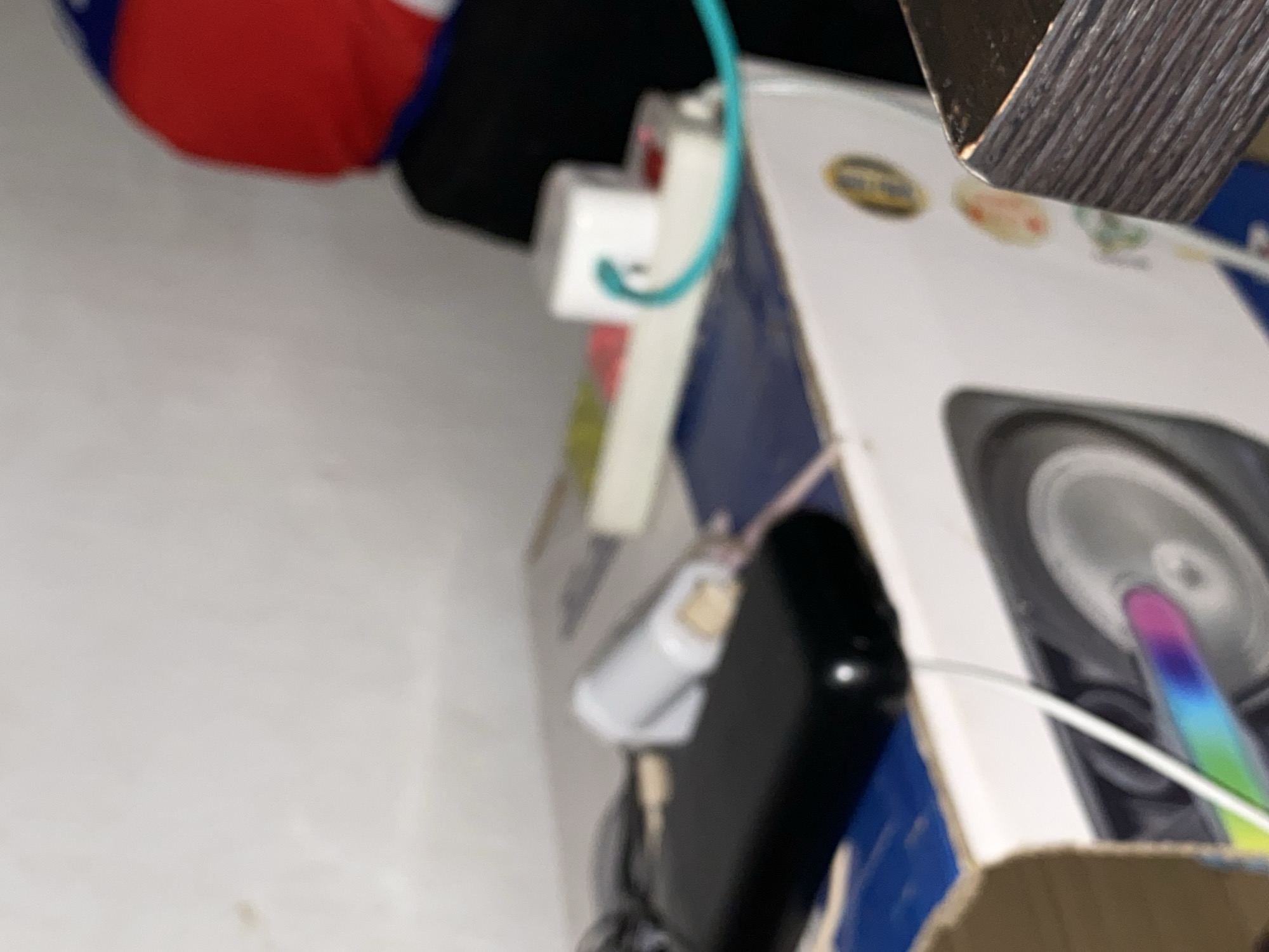 The image depicts a close-up, slightly blurred view of electronic devices and a power strip on top of a white box, possibly a computer tower or a large piece of equipment. A black object, likely a smartphone or portable charger, is in the foreground. A white cylindrical object with a wooden tip is plugged into the black device. Behind these, a white power strip is visible, with a white cube-shaped adapter plugged into one of its sockets. A turquoise cable snakes from the adapter and plugs into another socket on the power strip. The background shows a textured, off-white wall. In the upper right corner, a blurred object with red and blue colors suggests the presence of a person wearing a garment with these colors. The scene appears to be an indoor setting, possibly a room. There is no information available regarding the specific location in Jalingo, Nigeria.
