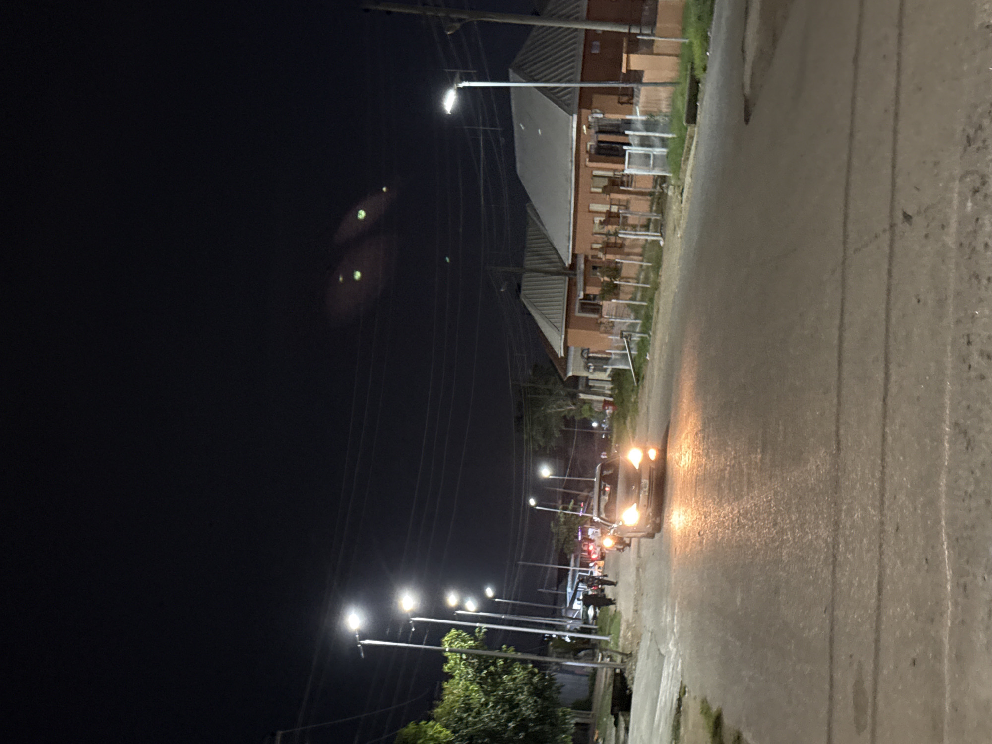 A nighttime, eye-level, long shot depicts a street in Abuja, Nigeria, illuminated by streetlights. The road is asphalt, wet from rain, and shows reflections of the lights. On the right side of the road, a row of residential buildings with orange walls and grey roofs is visible, separated by a fence. On the left side, a line of tall streetlights lines the road, with trees behind them. A silver sedan with its headlights on is driving towards the viewer in the center of the frame. Several people, silhouetted by the lights, are walking on the left side of the road. The sky is dark, with two bright, green, blurry lights that appear to be animal eyes reflecting in the distance.